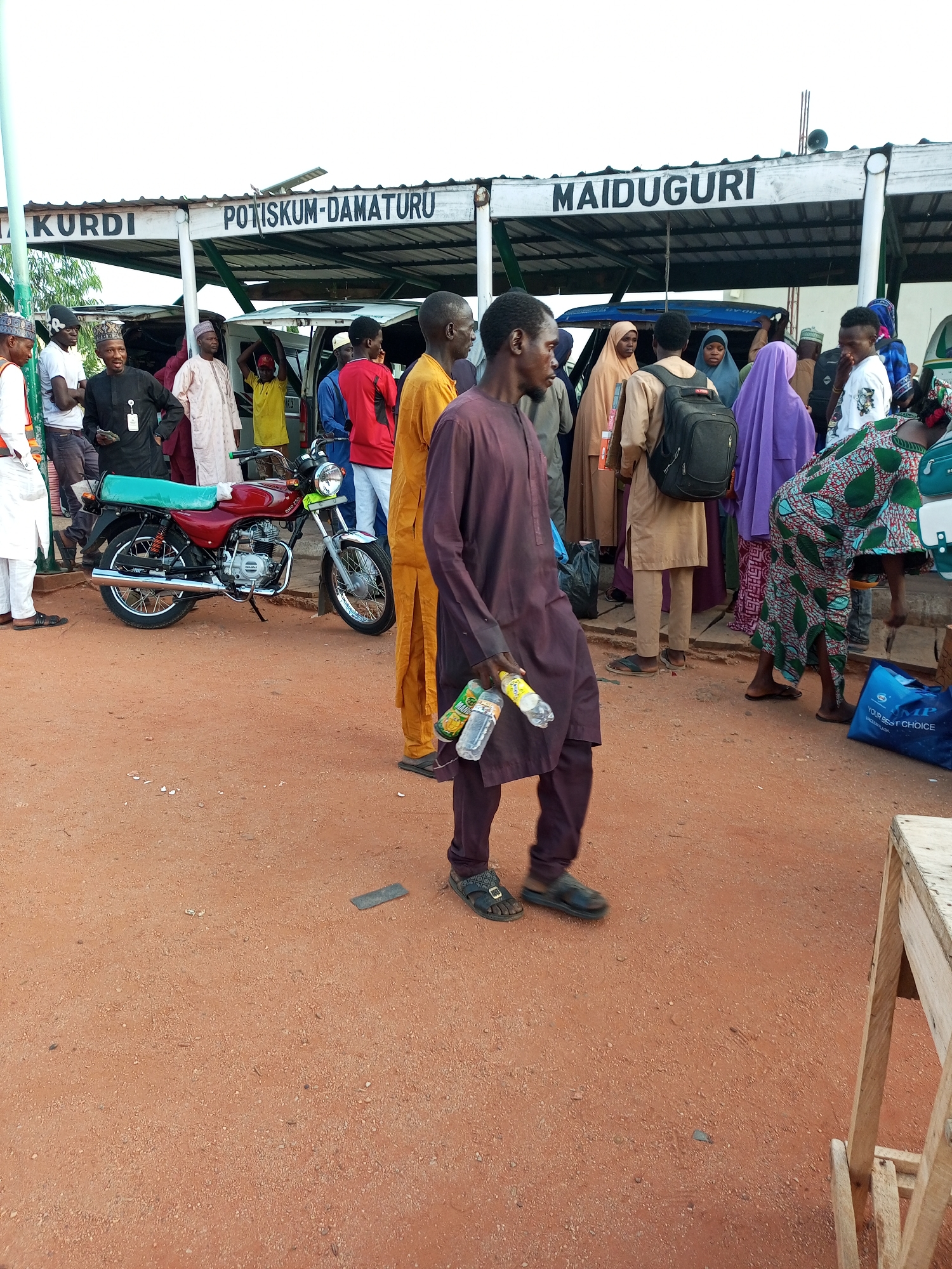 In Gombe, Nigeria, at what appears to be a transport hub, a group of people are gathered under a covered structure with signs indicating destinations: "KURDI", "POTISKUM-DAMATURU", and "MAIDUGURI". The ground is a reddish-brown dirt. In the foreground, a man in a deep purple outfit is walking towards the viewer, holding two plastic bottles of water. To his left, a red motorcycle is parked. Behind the man, various individuals are seen, some in traditional attire, others in more casual wear. A woman in a vibrant green and red patterned dress is visible to the right. Several blue plastic bags are on the ground. The atmosphere suggests a busy transportation area, possibly a bus or motorcycle park, with people waiting or preparing for journeys.