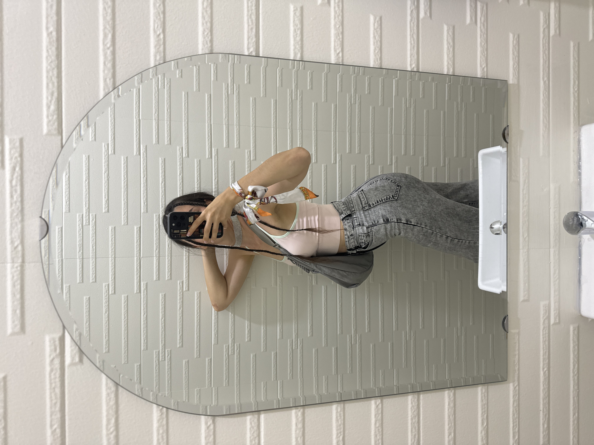 A woman is taking a picture of herself in a bathroom mirror. The mirror is tall and narrow with a curved top. The bathroom has white textured vertical tiles. The woman is wearing gray jeans, a light pink top, and a scarf around her wrist. She is holding a smartphone to take the picture. A white soap dish and other bathroom fixtures are visible on the right side of the mirror. The image appears to be from a location in Phu Thuy Ward, Vietnam.