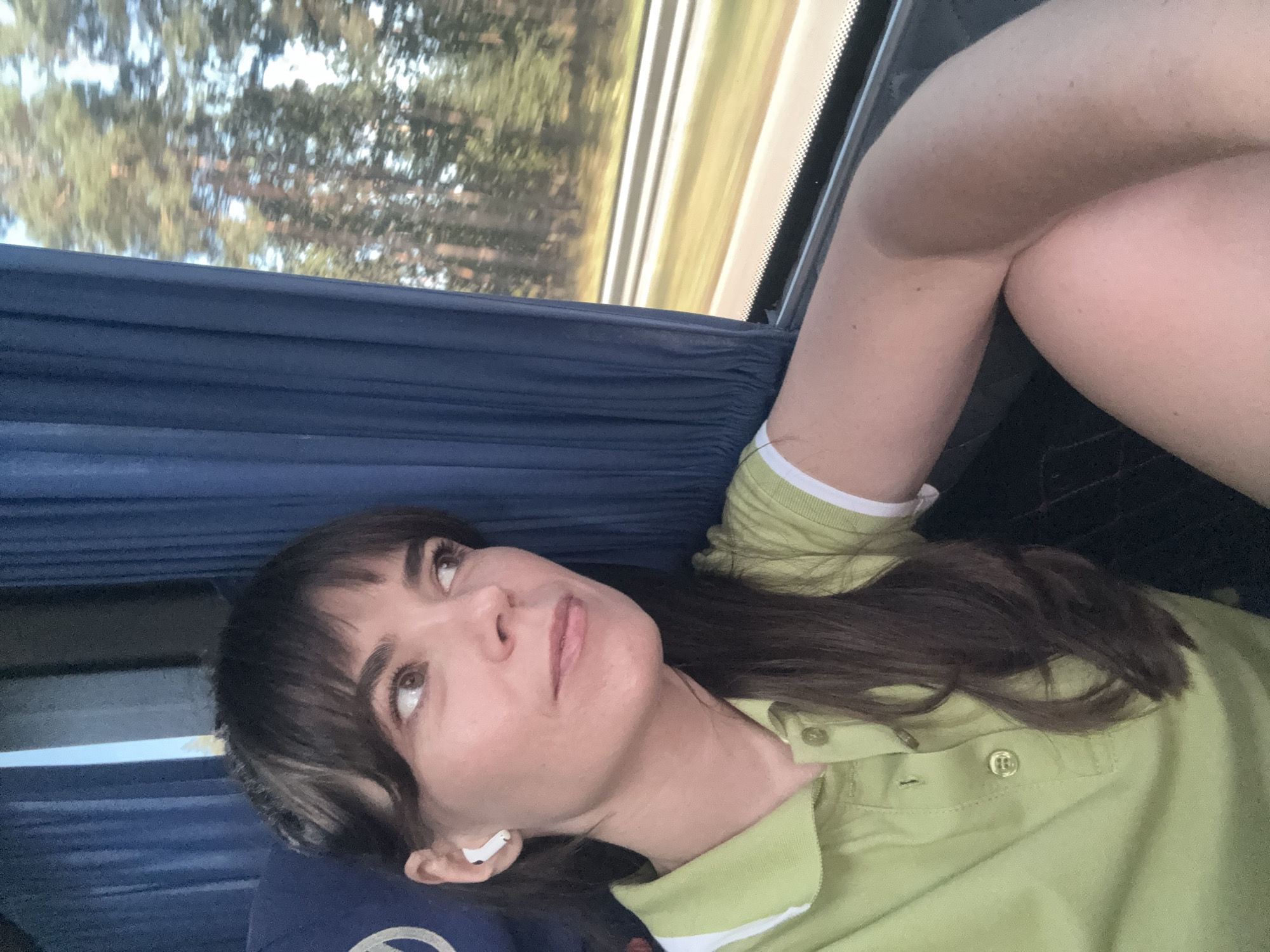 A woman with dark brown hair and bangs, wearing a light green polo shirt with white trim, sits in a vehicle. She is looking out the window, which shows a blurred view of trees and a road. A white wireless earbud is visible in her left ear. The interior of the vehicle appears to have blue curtains and a dark seat. The scene is captured from a selfie perspective.