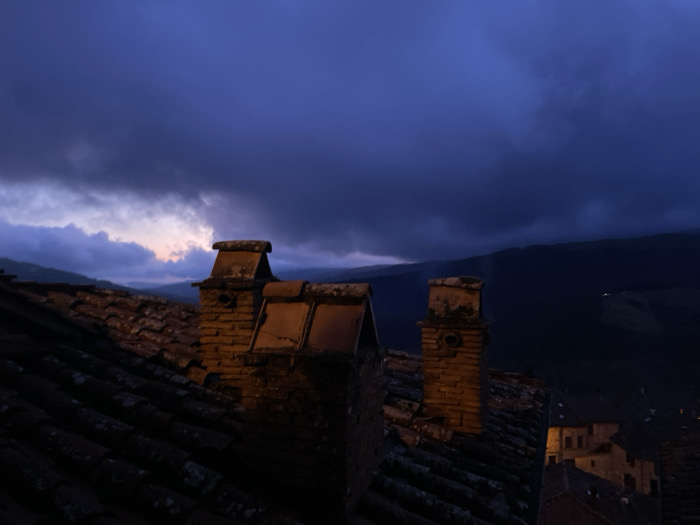 The image presents a somber yet evocative scene captured at dusk or night over the rooftops of Santa Fiora, Italy. In the foreground, several dark, aged terracotta tiled roofs slope downwards. Two prominent brick chimneys, crafted from warm-toned bricks and topped with stone caps, are illuminated by a soft, artificial light, casting a subtle glow on their textured surfaces. The weathered appearance of the tiles, possibly covered in lichen, suggests historical architecture.
Above the rooftops, the sky is dominated by heavy, dark blue and purple clouds, creating a dramatic and somewhat moody atmosphere. Towards the upper left horizon, a narrow band of lighter, almost pinkish-grey sky is visible beneath a thick layer of clouds, hinting at the last vestiges of sunset or the very beginning of dawn.
In the background, the landscape recedes into dark, rolling hills or mountains, largely obscured by the low light and dense cloud cover. Hints of distant structures with warm, glowing lights can be seen on the hillsides, further emphasizing the time of day. Below the main horizon, nestled among the rooftops, are glimpses of other traditional buildings with warmly lit windows, suggesting an old European town.
No people are visible, and no specific activity is taking place; the scene is static, focusing on the architectural and natural elements. The weather appears overcast and potentially damp, contributing to the quiet, introspective mood of the image. There is no visible text.