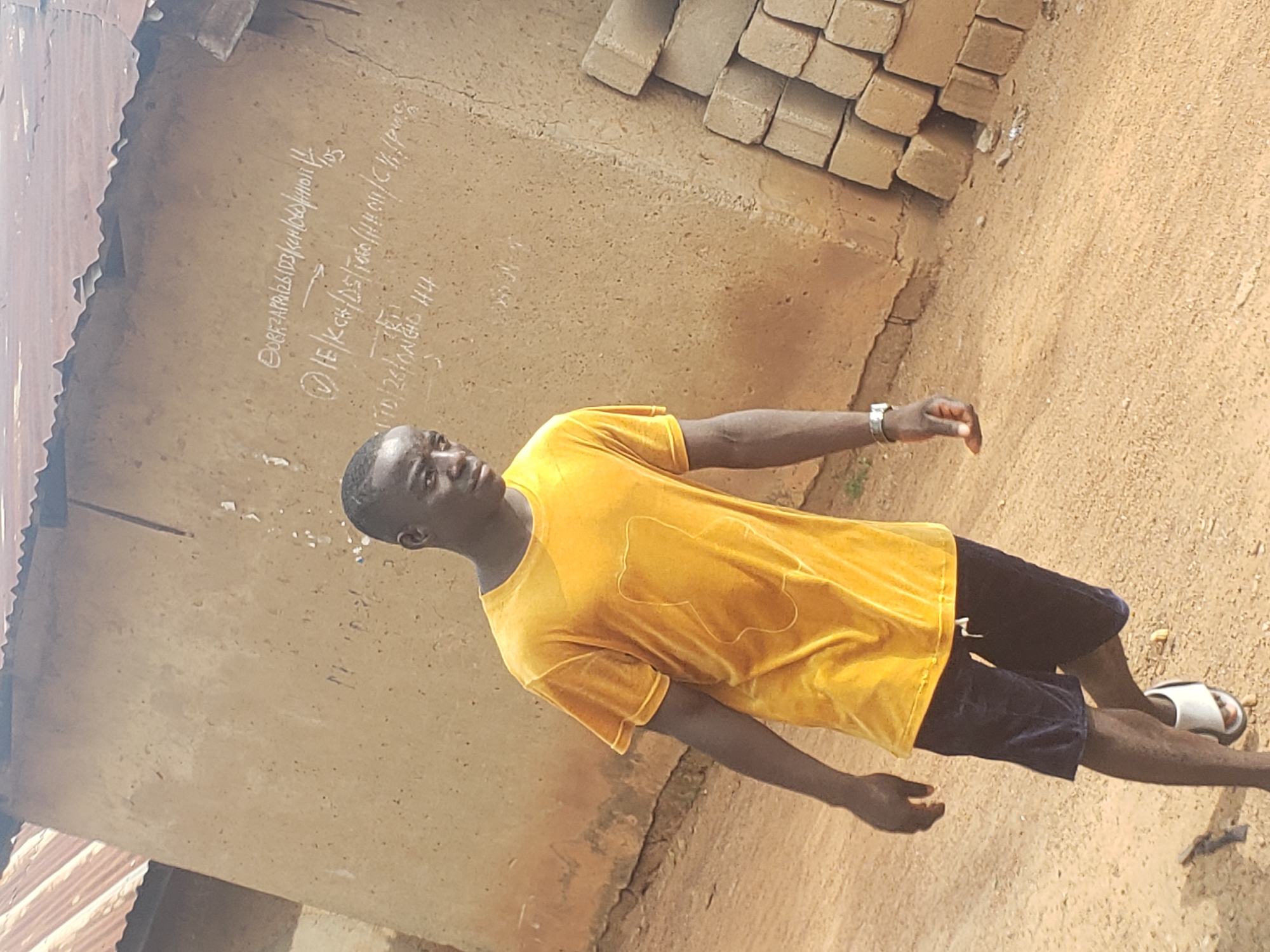A dark-skinned young man is walking in front of a clay building in Kachia, Nigeria. The man is wearing a bright yellow velour t-shirt and dark blue shorts. He has white flip-flops on his feet. The building has a corrugated metal roof and chalk writing on its walls. To the right of the man, there is a stack of bricks. The ground is dirt.
