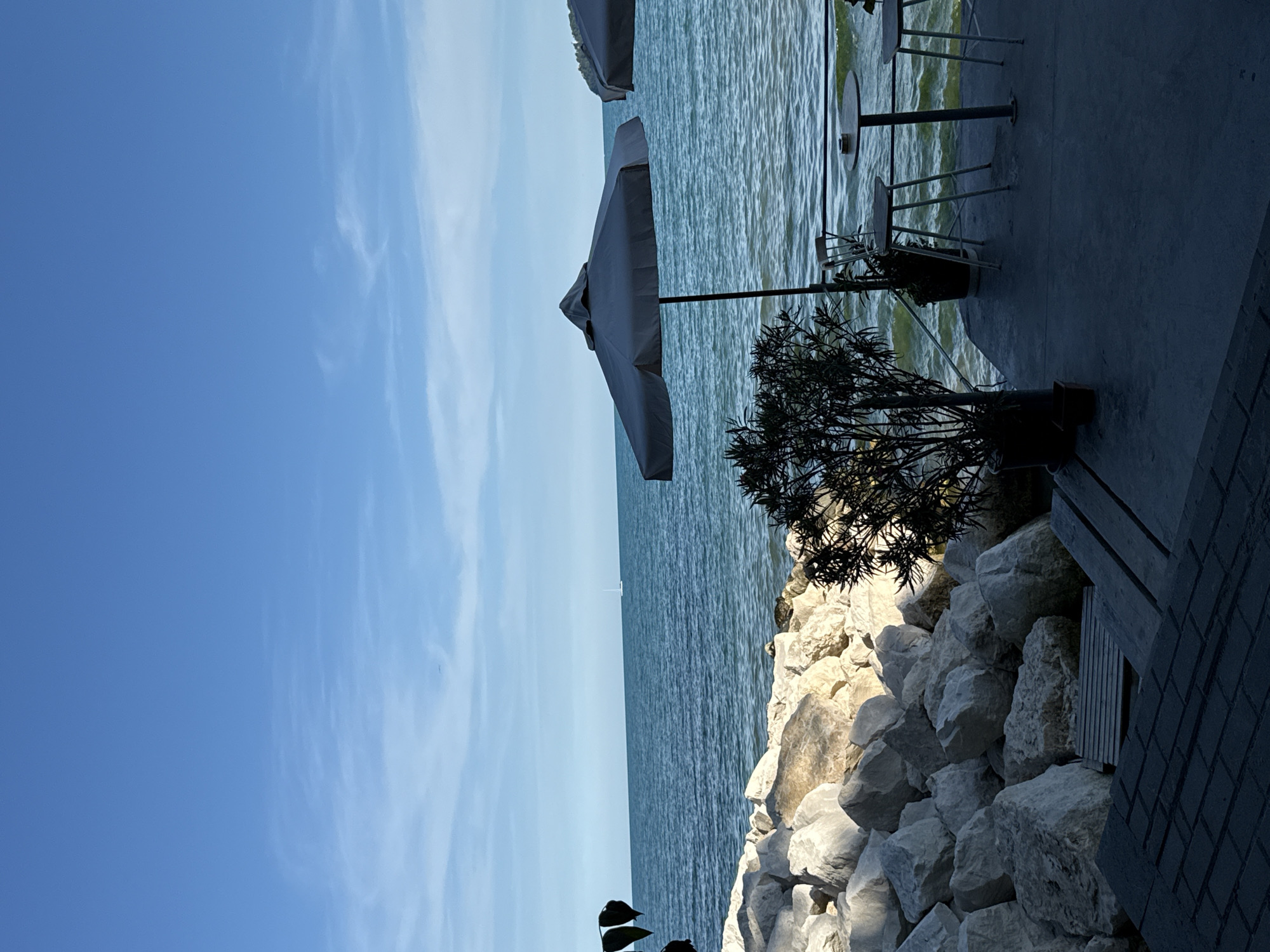 The image depicts an outdoor seating area adjacent to the sea, likely a cafe or restaurant terrace, with a rocky shoreline. The sky is a clear blue with sparse, wispy clouds. The sea is a deep blue-green, with moderate waves indicating a breezy condition. In the distance, a small sailboat is visible on the horizon.
The foreground shows a dark concrete patio. Two small tables and a few chairs are set up, with at least one umbrella providing shade. A potted plant with dark foliage stands near the tables. To the left, a substantial pile of large, light-colored rocks forms the edge of the land. A wooden bench or seating area is partially visible amongst the rocks.
The overall ambiance suggests a serene coastal location, possibly during the daytime. While the image does not provide explicit clues to pinpoint the exact location in Varna, Bulgaria, such seaside cafes with rocky shorelines are characteristic of certain areas along the Varna coast, particularly those with a more natural or less developed beach.