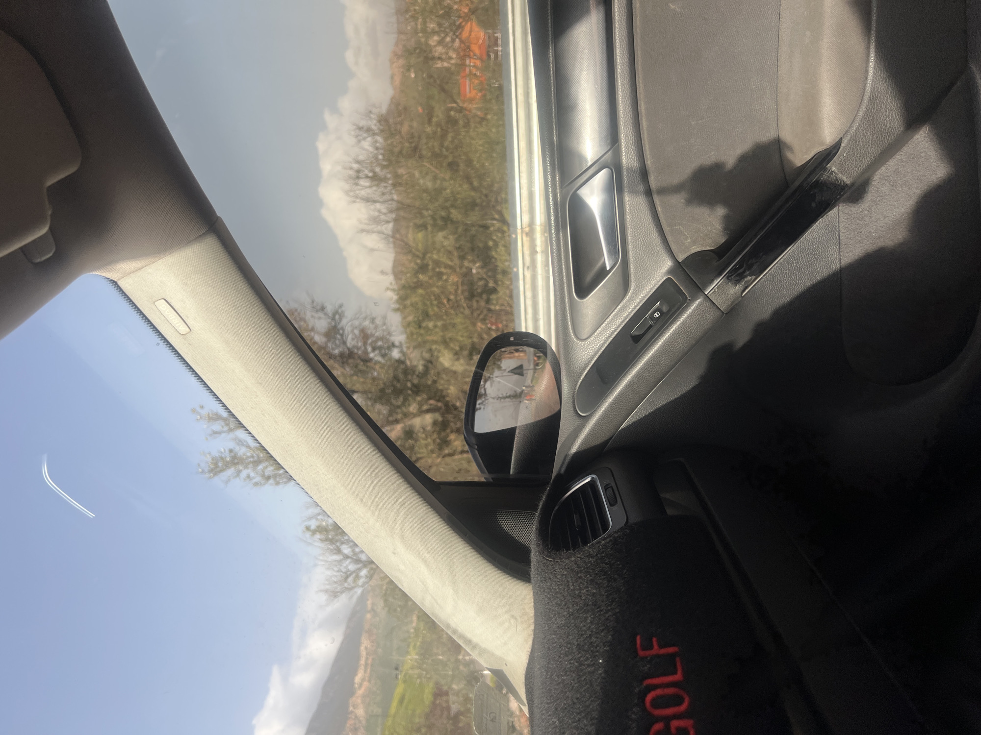 The image is taken from inside a Volkswagen Golf, showing the passenger side door, side mirror, dashboard, and part of the front windshield. The vehicle appears to be in Ouaouizert, Morocco, as evidenced by the reflection in the side mirror and the landscape visible through the windshield, which features mountainous terrain and sparse vegetation typical of the region. The car interior is dark, with a black dashboard and door panel. The door features a silver door handle and a lock/unlock switch. A black dashboard cover with the word "GOLF" stitched in red is visible near the air vent. The sky is blue with scattered clouds.
