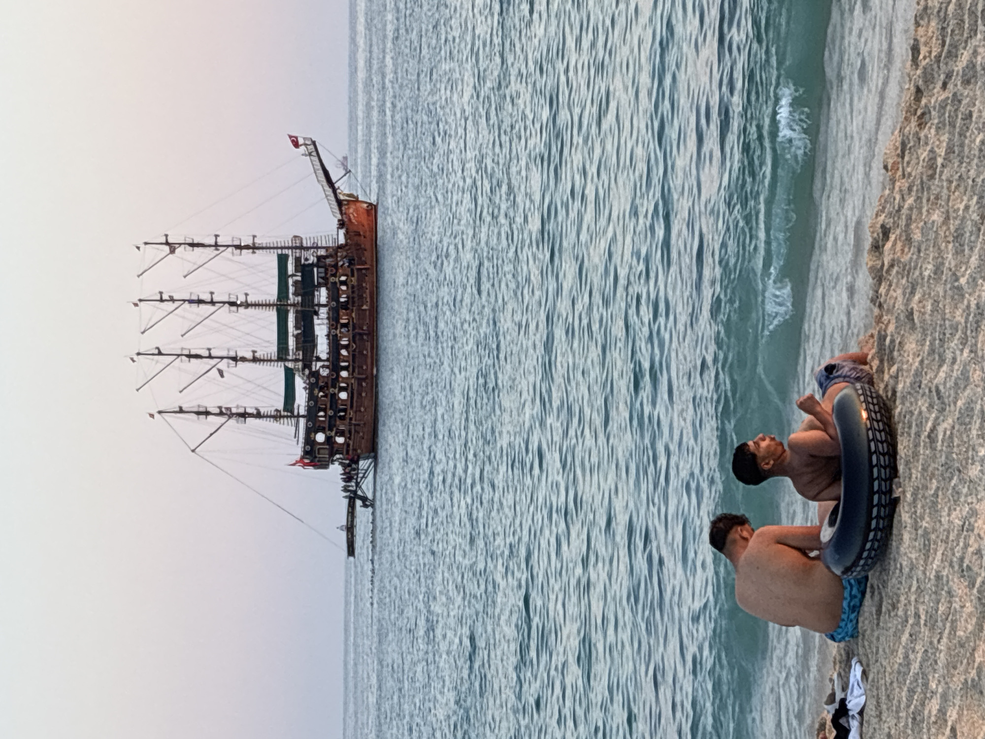 At the edge of the sandy shore in Alanya, Turkey, two young men are seated facing the turquoise sea. The man on the left is turned away from the camera, his back to the viewer, while the man on the right faces the sea, his body angled slightly to the side. He is positioned within a large, black inner tube. A few pieces of clothing lie on the sand to the left of the men. In the distance, a large, multi-masted wooden ship, reminiscent of a pirate ship, sails on the calm water. The ship has several decks, numerous windows, and is adorned with flags. The sky above is a pale, gradient hue, transitioning from a light pinkish-orange near the horizon to a soft grey at the top. The overall scene depicts a serene coastal environment at dusk.