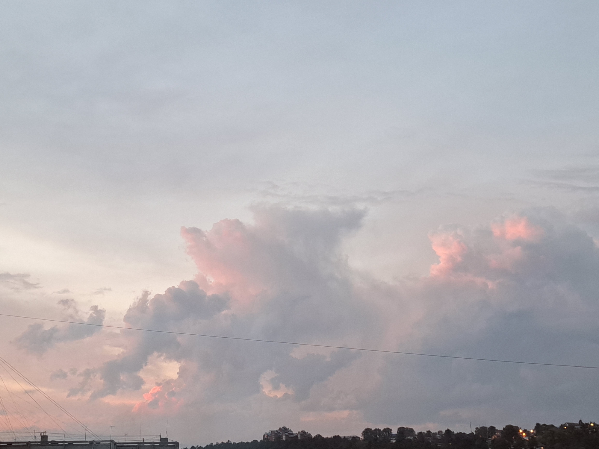 The image shows a cloudy sky at dusk with pink and gray clouds. A thin black wire stretches horizontally across the middle of the frame. Below the clouds, a silhouetted treeline with a few buildings is visible. The overall impression is a peaceful, twilight sky. There are no people or specific landmarks that would indicate the location as Irkutsk, Russia.