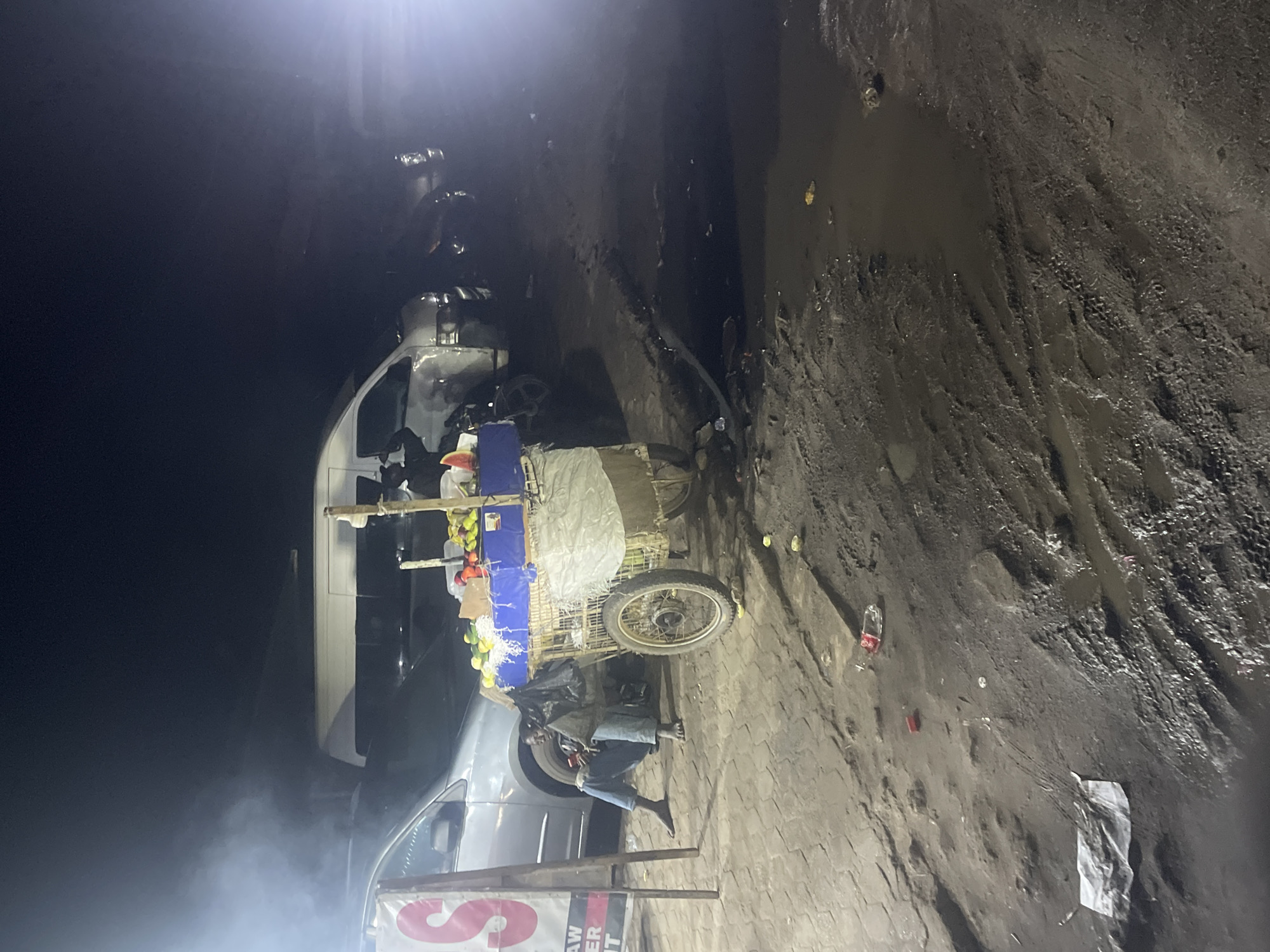 A street scene in Obiaruku, Nigeria at night. A man sits on a low seat beside a fruit cart filled with produce. The cart stands next to a light-colored van. The ground is uneven and muddy in places, strewn with litter. A sign is visible in the foreground, partly obscured by smoke or fog. The scene is dimly lit.