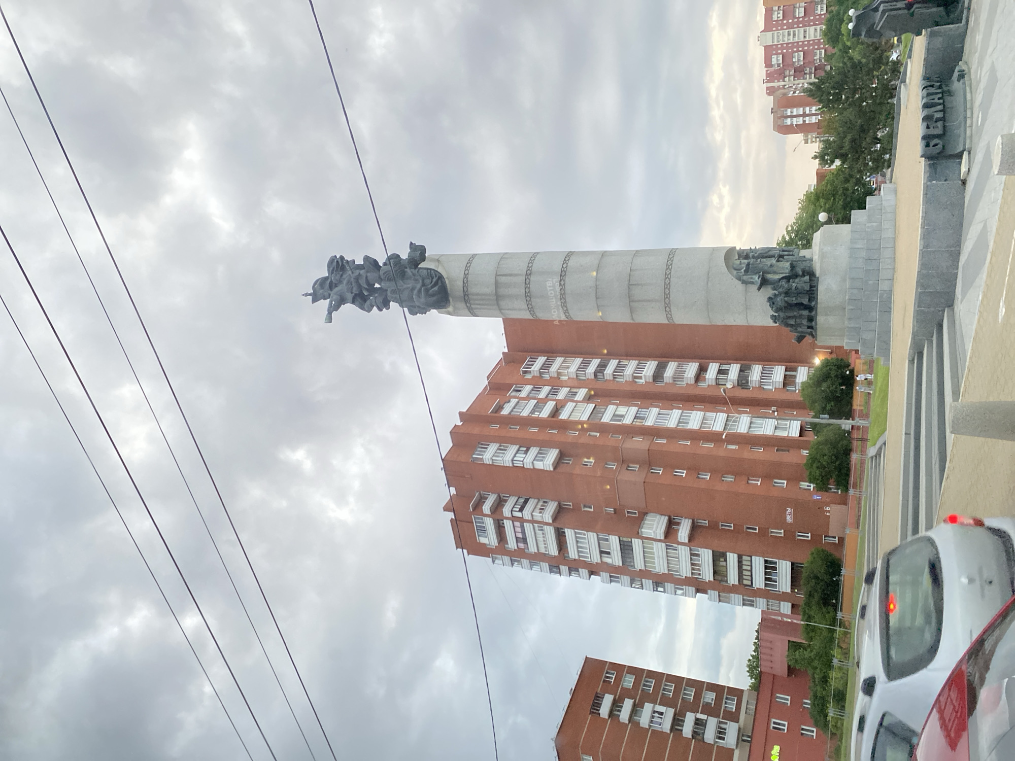 The image depicts a monument with figures on top of a tall, cylindrical structure. The monument is situated in an urban setting, with a tall, red brick apartment building visible behind it. In the foreground, there are power lines, and a white car is partially visible. The scene appears to be outdoors, possibly in a public square or park, given the presence of the monument. The sky is overcast, suggesting it could be daytime with cloudy weather. The overall atmosphere is somewhat solemn, likely due to the nature of monuments which often commemorate historical events or figures. The text "ЦВЕТЫ" (flowers) is visible on a sign on the building. The location is identified as Лугаваслабадскі сельскі Савет, Belarus.