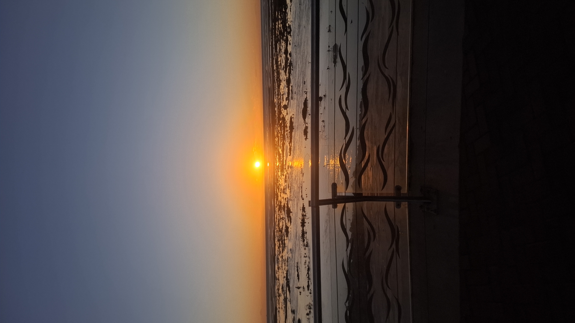 The image captures a vibrant sunset or sunrise over a vast body of water, likely the Río de la Plata in Villa Domínico, Argentina. The scene is dominated by a bright, intensely orange-yellow sun positioned low on the horizon, casting a strong glare that reflects across the water's surface.
The sky transitions from a deep, clear blue-grey at the top of the frame to warm hues of orange and yellow closer to the horizon, indicative of either dawn or dusk. The expansive body of water, appearing calm, shows shimmering reflections of the sun's light.
On the right side of the photograph, a wooden or panelled structure, possibly a wall, fence, or part of a building, partially frames the view. This structure features decorative, dark, wavy patterns carved or painted onto its surface. A metallic latch or handle is visible on this structure in the lower right portion of the image. The sunlight casts warm highlights on the texture and patterns of this architectural element.
There are no people or discernible text visible in the image. The overall impression is one of natural beauty and serenity during a clear and colorful sunset or sunrise.