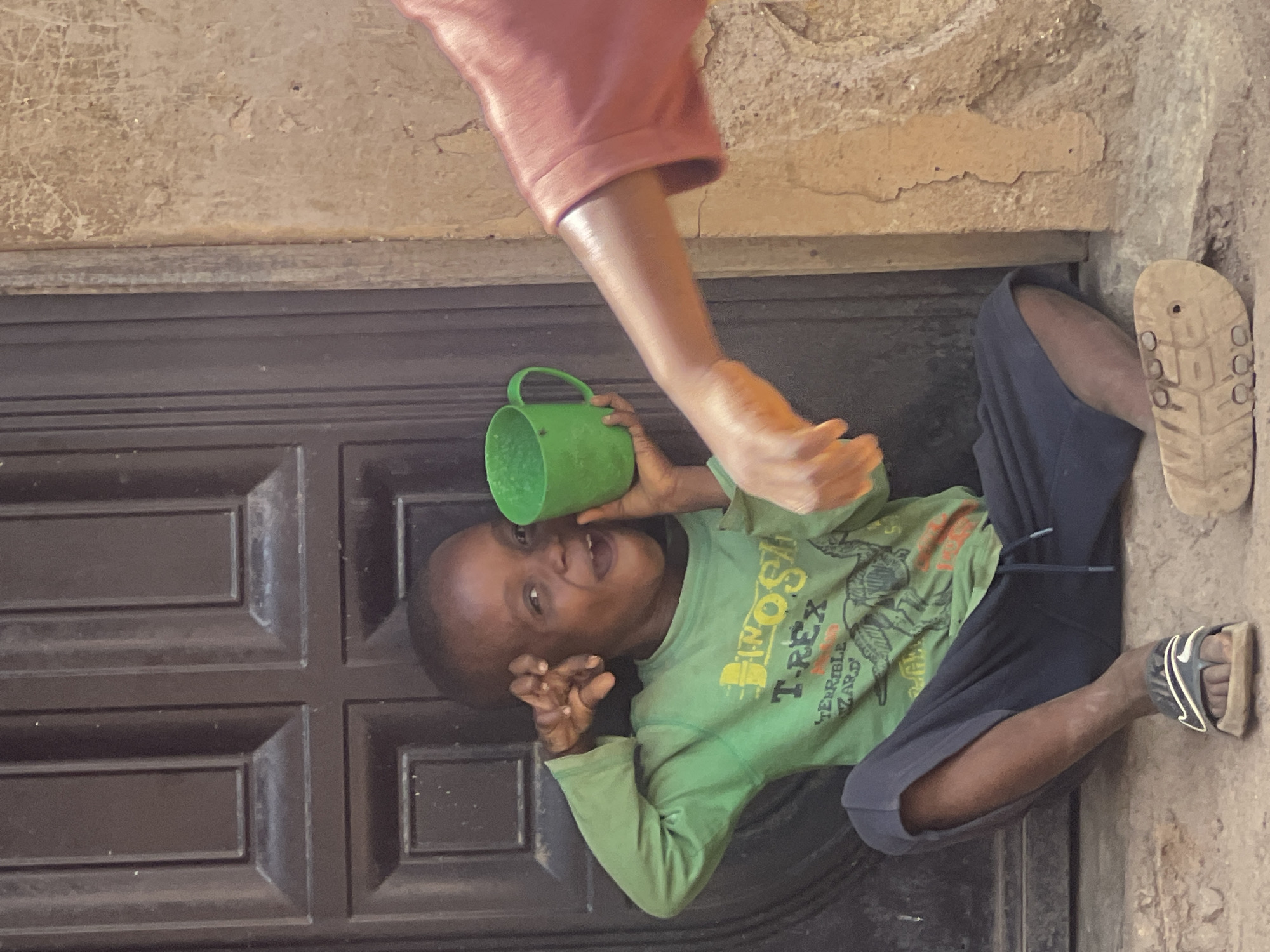 A child in a green shirt and black pants sits in front of a dark brown door, holding a green cup to his ear. An adult's arm in a pink sleeve is extended toward the child. A beige sandal lies on the ground near the child's feet, alongside a black and white sandal worn on the child's foot. The scene appears to be located in Gura Top, Nigeria, based on contextual information.