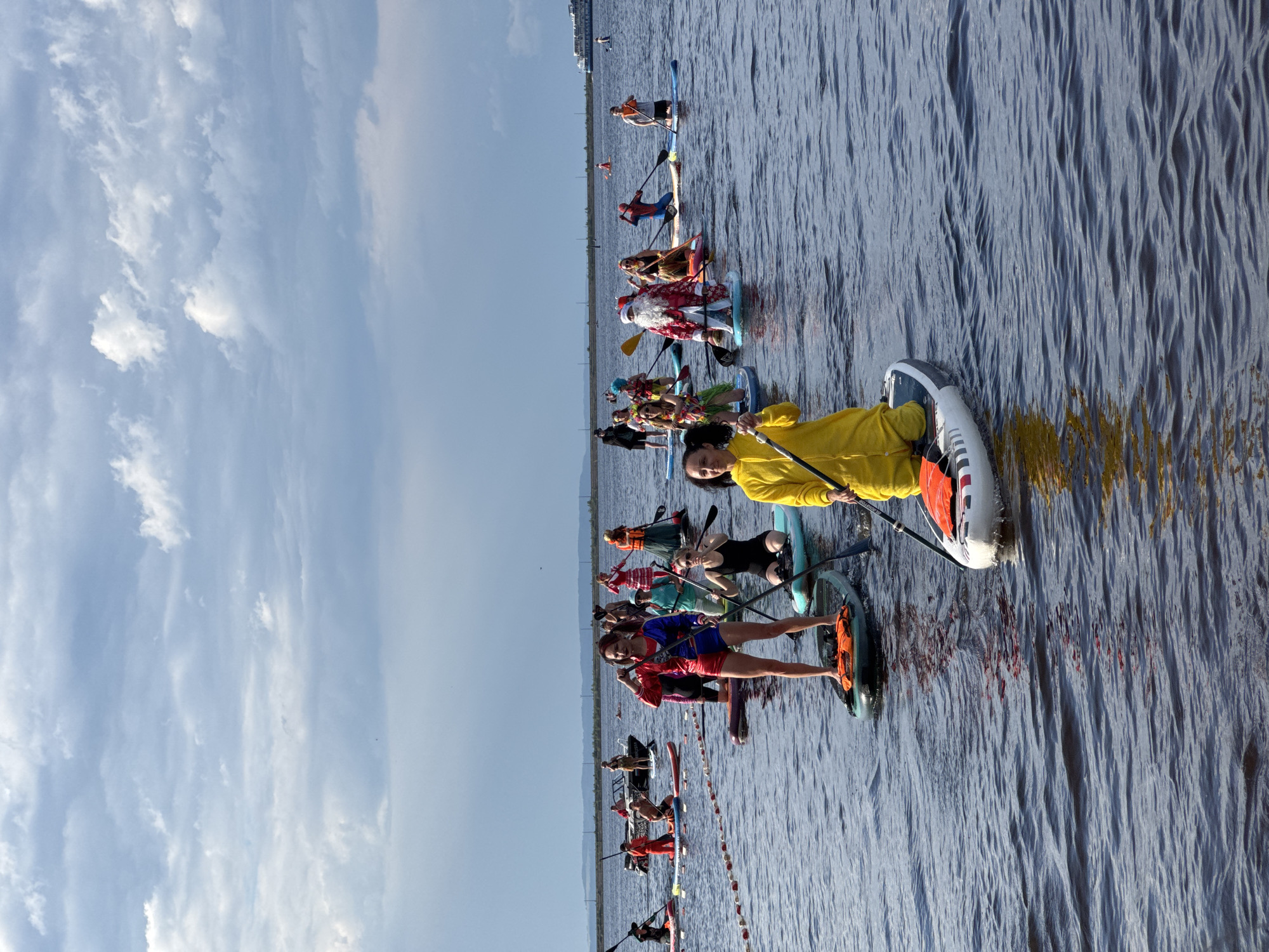 A group of people in costumes are paddleboarding on a body of water. Some of the costumes appear to be festive, with one person dressed as Santa Claus and others in bright, colorful outfits. The water is relatively calm, and the sky is partly cloudy. In the background, there are some distant landmasses and what appears to be a ship or ferry. The setting is likely a lake or a wide river, potentially near a coastline given the presence of the vessel in the distance. The event seems to be a recreational gathering or possibly a themed event in Chaikovsky, Russia, given the prompt's reference to the city. The focus is on the individuals on their paddleboards, engaged in the activity.