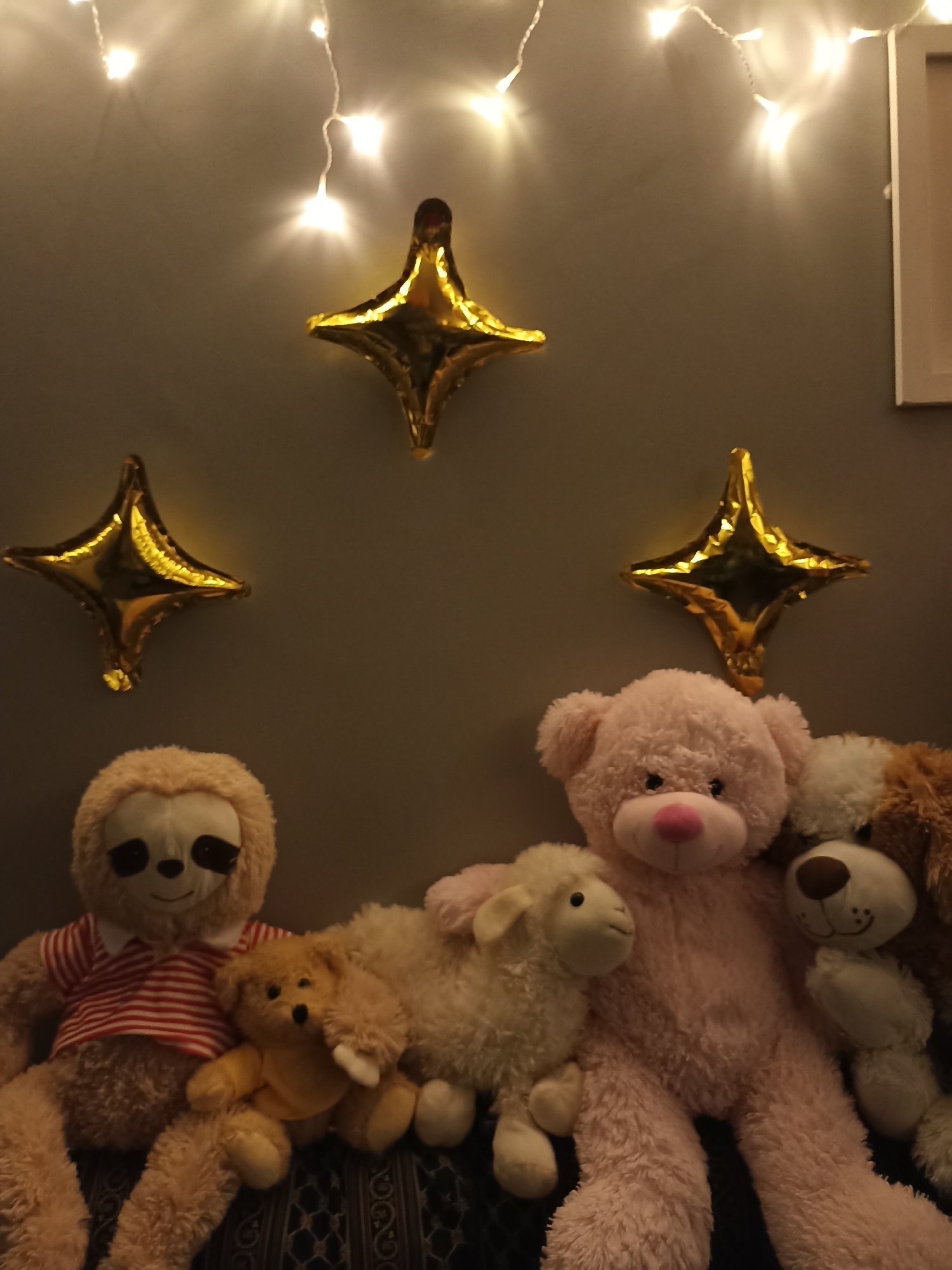 The image shows a collection of plush toys arranged on a surface in front of a grey wall. String lights with a warm glow are visible at the top of the image, and three gold star-shaped balloons are suspended on the wall behind the toys.
The plush toys, from left to right, include:
* A large tan sloth wearing a red and white striped shirt.
* A small tan bear wearing a yellow garment.
* A white sheep.
* A large pink teddy bear.
* A brown and white dog with floppy ears.
The toys are positioned closely together, appearing to be set up for display or a photograph. The background is a plain grey wall with the decorative elements of lights and balloons. The setting does not provide any specific clues to its location within Annagry, Ireland.