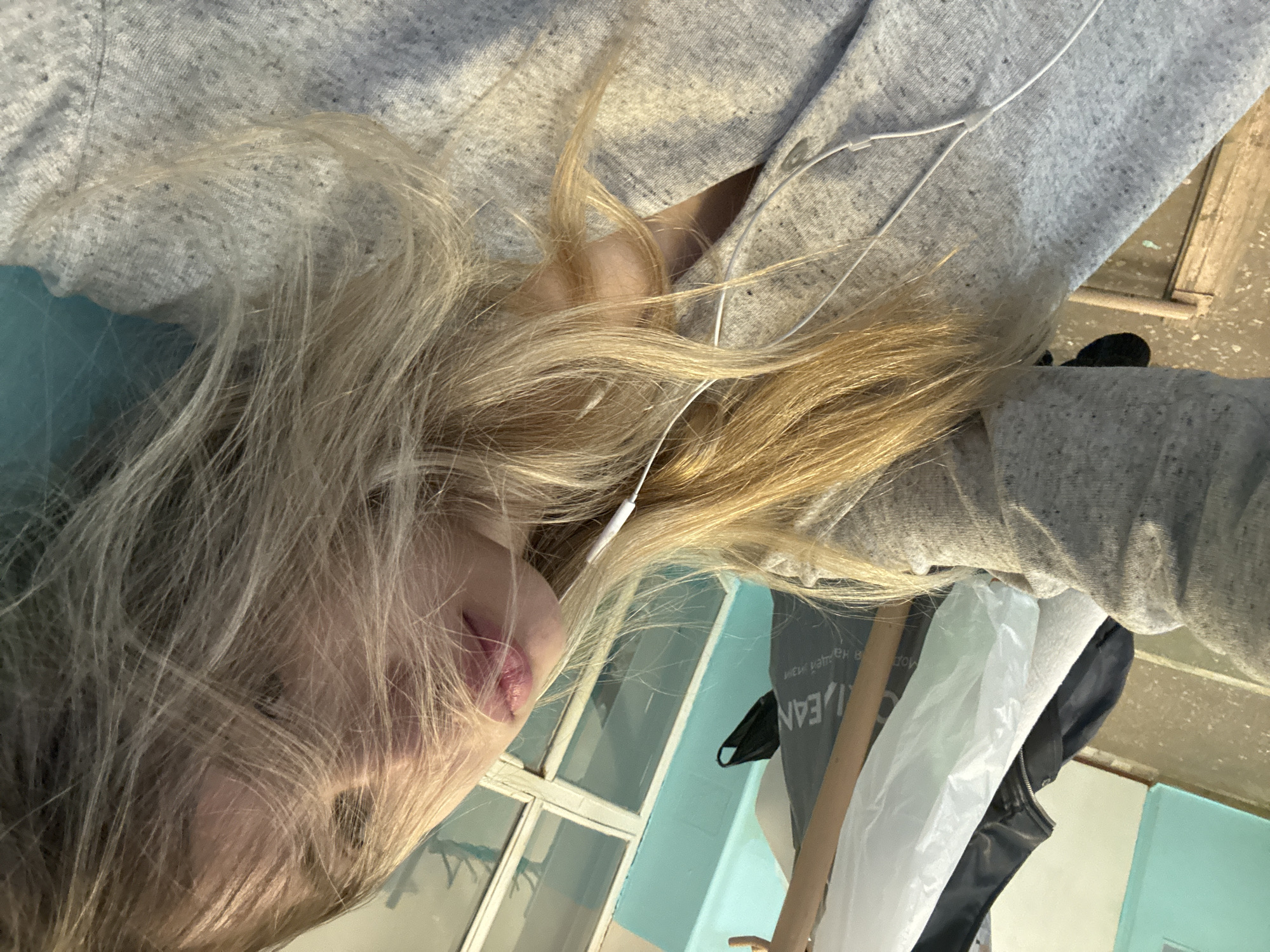 A young woman with blonde hair is lying down, wearing a gray sweatshirt and white earphones. Her face is visible, and she appears to be looking slightly upward. A window with a teal frame and white panes is visible in the background. A black face mask hangs from a gray bag. Part of a white plastic bag and another black jacket are also visible. The location is not identifiable based on the image.