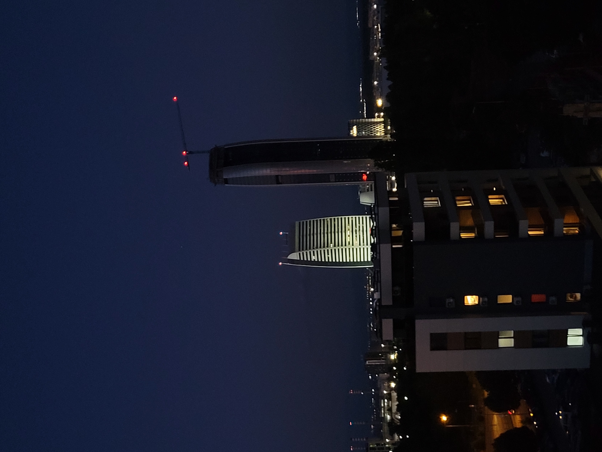 This is a nighttime, eye-level, medium shot of the Sofia skyline in Bulgaria, featuring modern architecture. In the foreground, a dark, multi-story building with illuminated windows is visible, its facade predominantly gray and white. To the left of the foreground building, a street lamp with a yellow light illuminates a portion of a road and some dark trees.
In the mid-ground, two prominent skyscrapers dominate the scene. The left skyscraper is characterized by its distinctive white, horizontal striped facade and is brightly lit from within. The right skyscraper is a tall, dark, and sleek tower, partially under construction, indicated by a construction crane at its apex with three red lights. To the right of this skyscraper, another building with a patterned facade is illuminated.
The background reveals a dense urban landscape with numerous buildings and lights, suggesting a sprawling city. A dark, tree-lined area occupies the lower right portion of the image, separating the foreground and mid-ground buildings from the distant cityscape. The sky is a deep, dark blue, characteristic of nighttime. A single, small, distant light can be seen in the upper right quadrant of the sky.