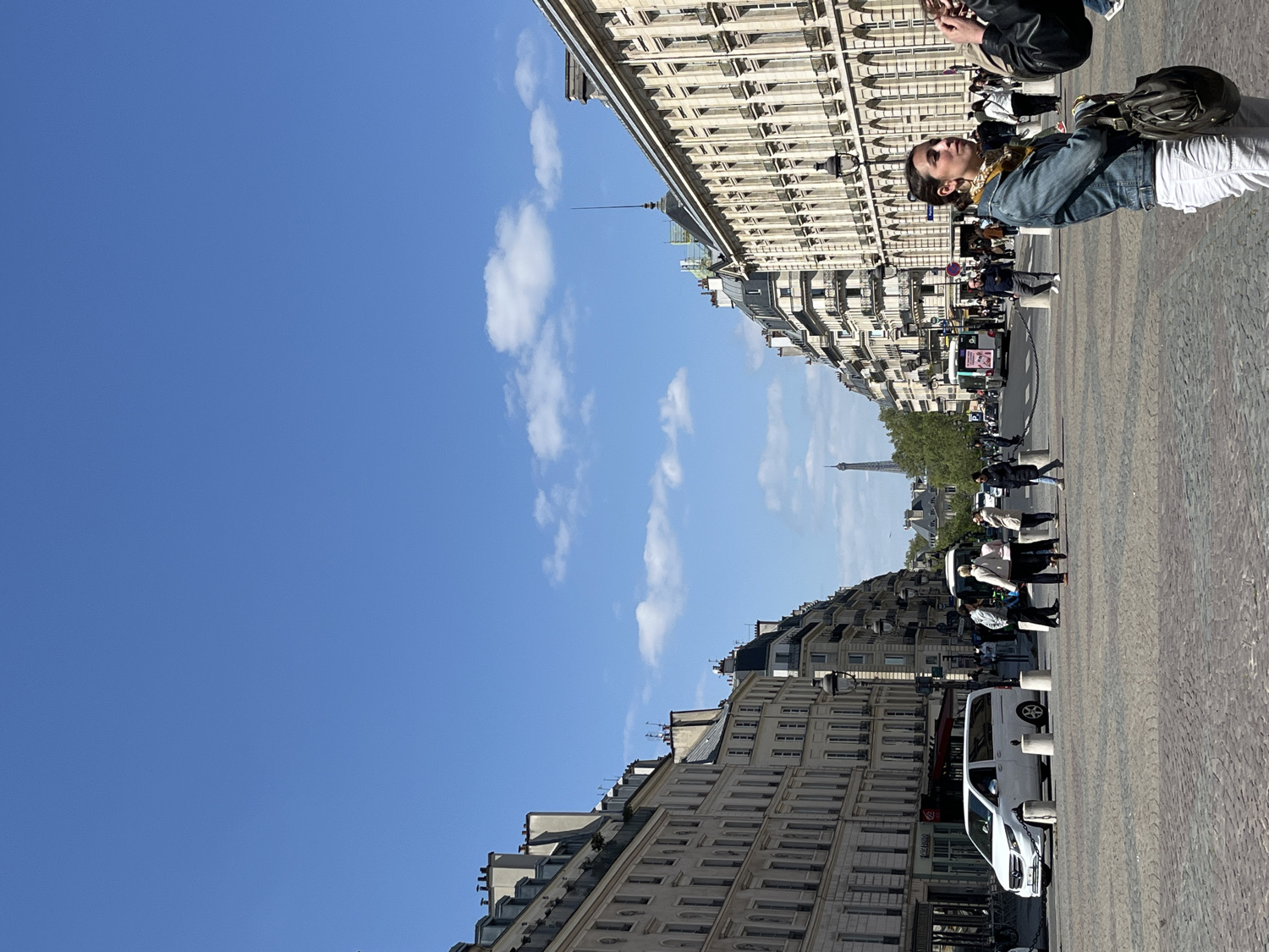 The image shows a Parisian street on a clear day with the Eiffel Tower visible in the distance. The street is lined with buildings, and there are people walking along the sidewalk. A person is looking at the Eiffel Tower. A white van is parked on the street. The sky is blue with some clouds. The street is paved with cobblestones.