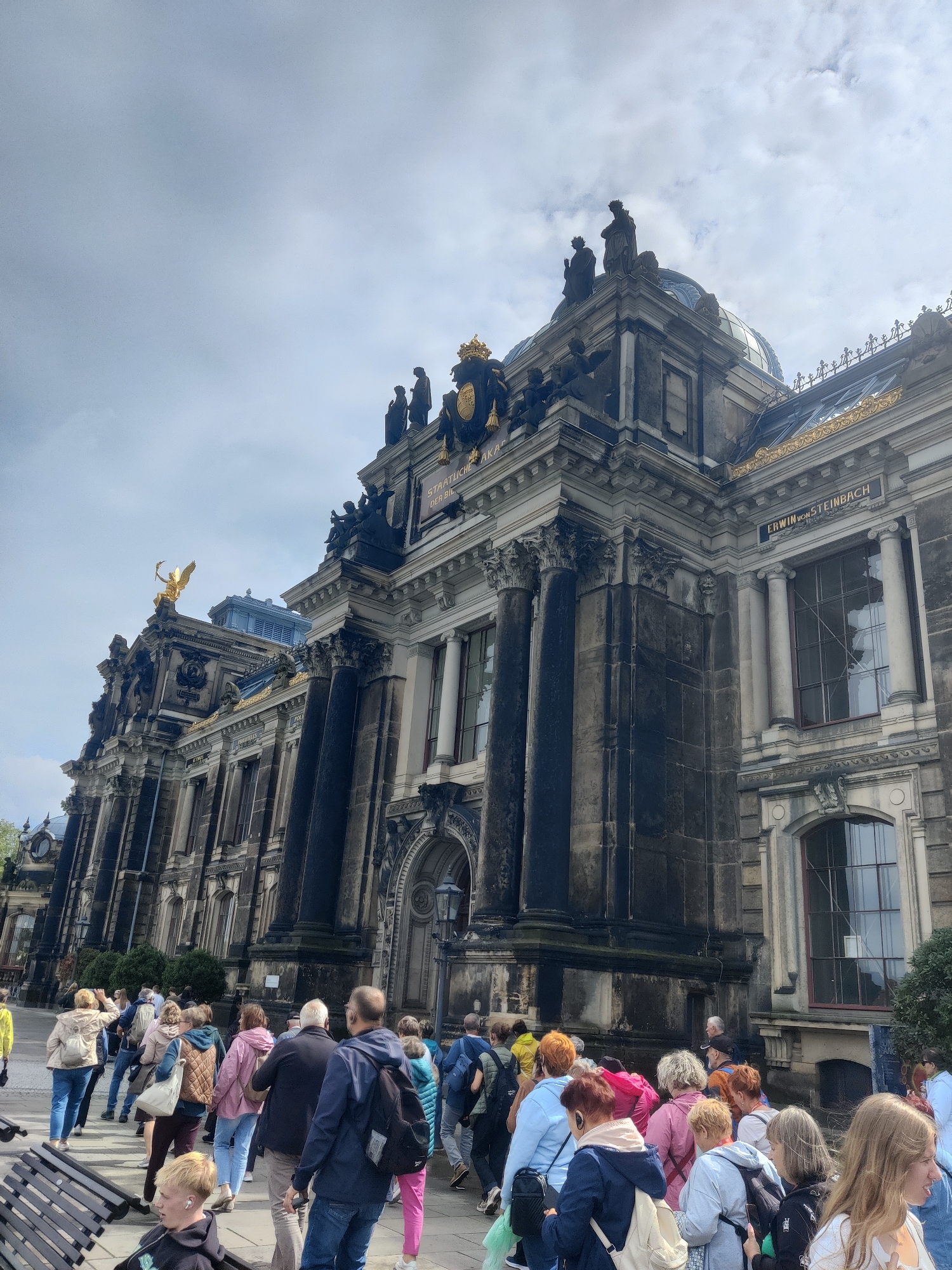 This is an eye-level, full shot image capturing a group of tourists walking towards a grand, ornate building in Dresden, Germany. The building is a large, dark stone structure with classical architectural elements, including columns, statues, and gilded embellishments. The sky above is cloudy, with a mix of grey and white.
In the foreground, several people are walking away from the camera, towards the building's entrance. They are dressed in casual clothing, with backpacks and purses indicating they are likely on a tour. Some individuals are looking towards the building, while others are engaged in conversation. A dark wooden bench is visible in the lower left corner of the frame, with a young man sitting on it, facing to the left.
The building itself is the main subject, with its intricate details drawing the viewer's attention. There are multiple levels, with large windows and decorative statues adorning the facade. A prominent golden statue of a winged figure is visible on the left side of the building, and lettering on the facade reads "STAATLICHEN AKADEMIE" and "ERWIN VON STEINBACH". The overall impression is one of historical grandeur and cultural significance. The setting appears to be a public area, possibly a plaza or walkway in front of the building.