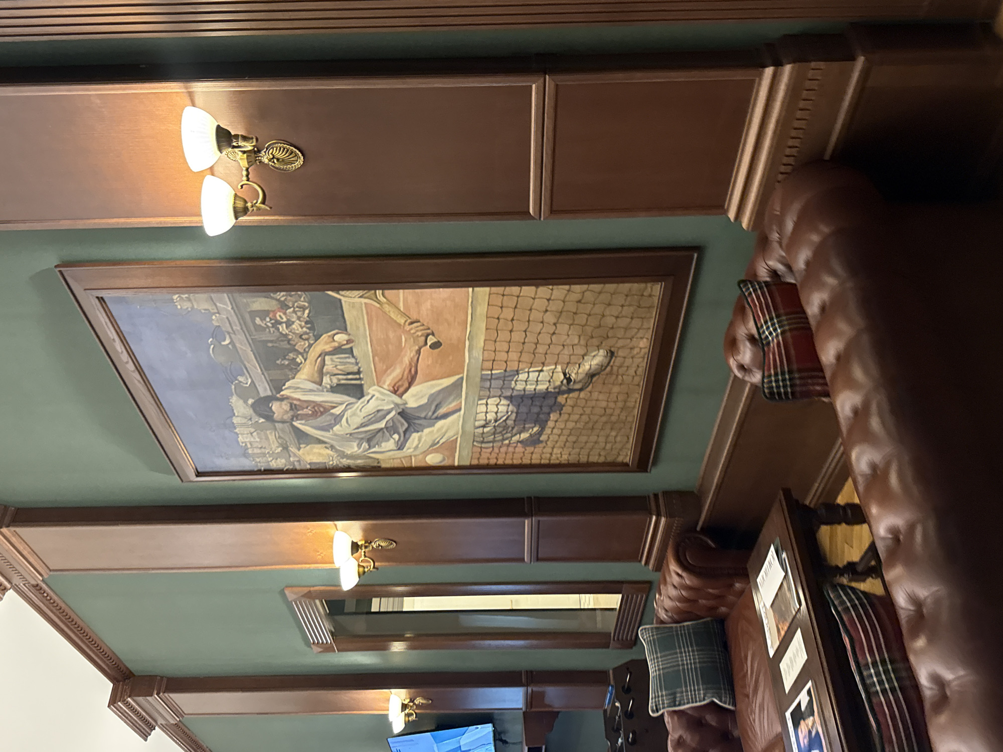 The image depicts an indoor scene, likely a lounge or club, with dark wood paneling on the walls and ceiling. Dominating the space is a large painting of a tennis player in mid-action, serving a ball. The painting is framed by dark wood and hangs on a teal-colored wall. Above the painting, a pair of brass sconces with white glass shades illuminate the area.
To the right of the painting, a plush, tufted leather sofa is visible, adorned with plaid cushions in red and green hues. Another section of the same sofa is seen further to the right, showing a tufted texture and dark brown leather. A low wooden table sits in front of this sofa, displaying several magazines and what appear to be framed photographs or prints.
The lighting suggests it might be evening or an interior space without natural light. The overall ambiance appears to be one of relaxed sophistication, possibly a place for leisure and conversation. The presence of the painting and the comfortable seating arrangement indicate a setting designed for enjoyment and respite. There is no visible text in the image.