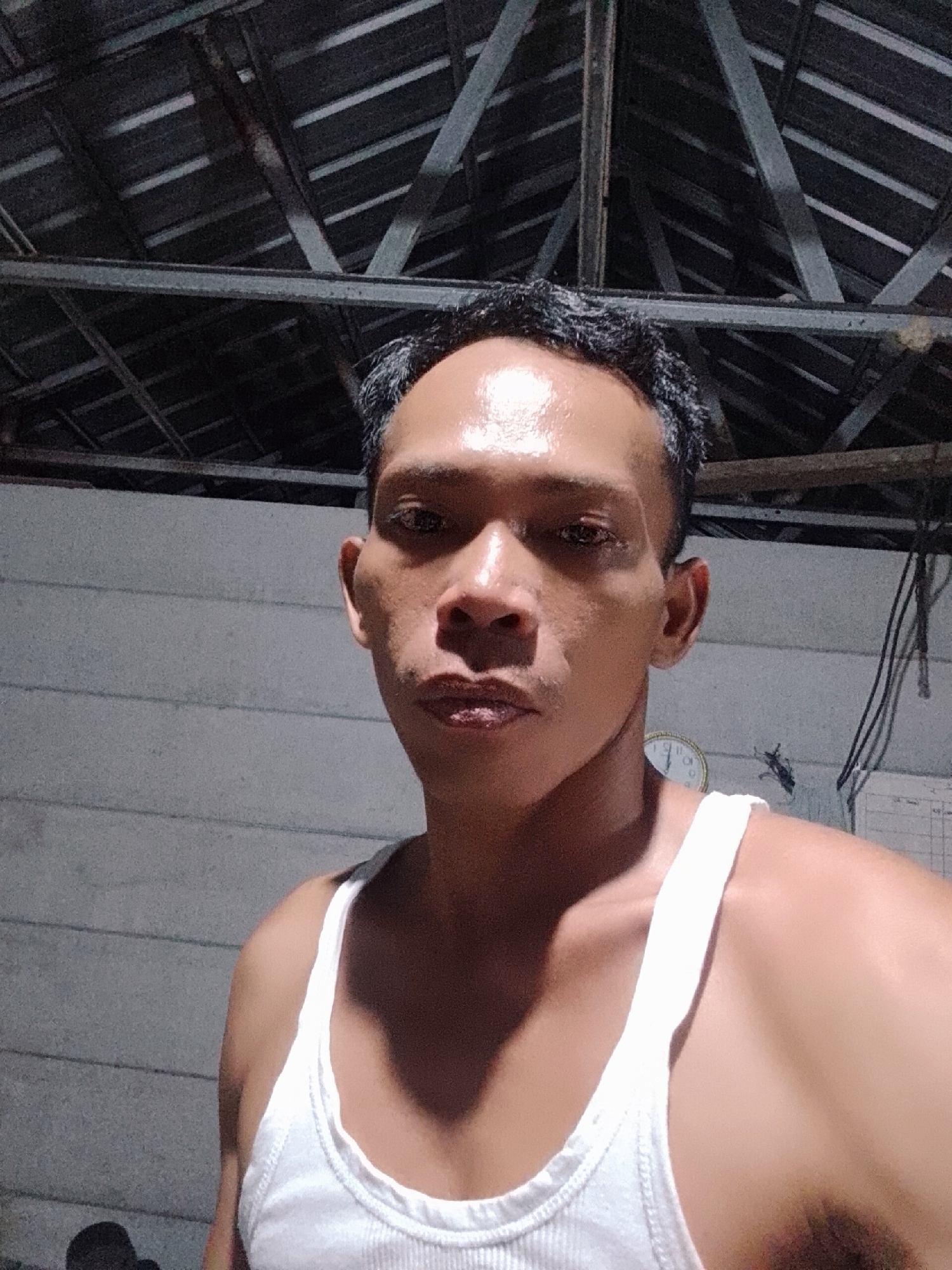 A medium-shot photograph depicts a middle-aged man in a white tank top. The man has short, dark hair, and a serious expression. The background includes corrugated roofing with exposed framing, a wall, an analog clock, and wires. The location is suggested to be Bukit Ketok, Indonesia.