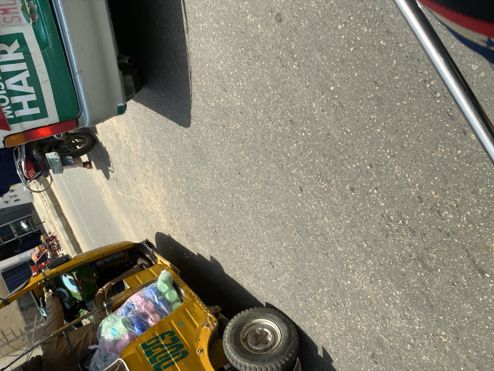 This image shows a street scene in Sabon Gari East, Nigeria. The primary subjects are two vehicles: a silver and green vehicle with "MOISTURIZING HAIR" written on its side and a yellow auto-rickshaw loaded with plastic bottles. The auto-rickshaw is positioned in the foreground, and a person is visible inside, seemingly driving or operating it. The other vehicle is partially visible in the upper left, with its rear end angled towards the road.
The scene appears to be a street with an asphalt surface. There are shadows cast on the road, suggesting it is daytime, likely mid-morning or afternoon. The overall impression is one of daily commerce or transportation. The auto-rickshaw is carrying a load of colorful plastic bottles, some in green, pink, and blue, wrapped in clear plastic. The silver vehicle has a license plate visible, though the text is not clearly legible.
The image is oriented vertically, with the road occupying a significant portion of the frame. The presence of the auto-rickshaw, a common mode of transport in many parts of Africa and Asia, along with the style of the vehicles and the general street appearance, aligns with the provided location context of Sabon Gari East, Nigeria. No specific emotions are conveyed, but the image depicts a typical urban or semi-urban environment with active transportation.