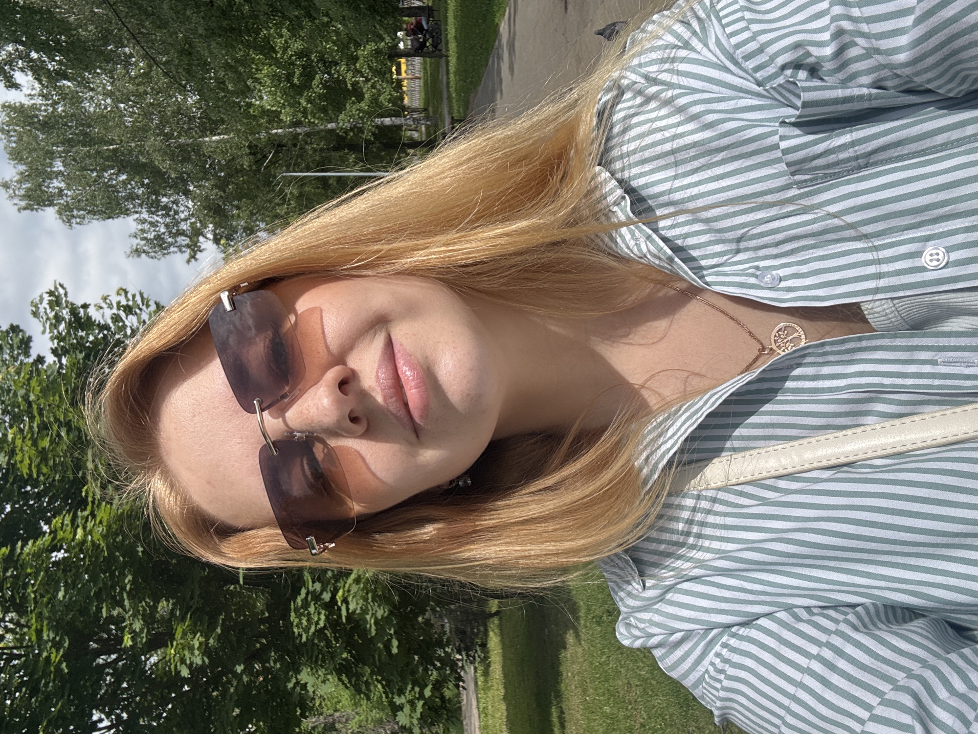 The image is a close-up, slightly tilted selfie of a person outdoors in a park in Podosenikha, Russia. The subject is a female with long, blonde hair, seen from the chest up. She is wearing large, dark brown-tinted sunglasses with a frameless design. Her lips have a subtle, natural pink hue, and she displays a relaxed, slight smile. She wears a light-colored shirt with thin horizontal stripes, likely in a pale green or mint shade, with visible buttons down the front. A delicate golden chain necklace with a circular "tree of life" pendant hangs around her neck. A cream or light-colored strap from a shoulder bag is visible across her chest.
The setting is a sunny outdoor environment, characterized by abundant green trees and grass, suggesting a park or garden. A paved path is visible in the background, along with distant elements that appear to be park benches and possibly other individuals, though they are out of focus. The bright sunlight creates clear shadows on her face and clothing, indicating daytime, likely late morning or afternoon. The sky, visible through the trees in parts, appears bright with some clouds. There is no visible text in the image.