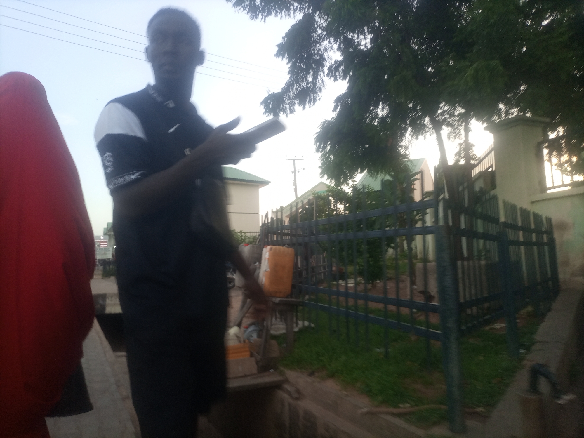 The image shows an outdoor scene in Arawa, Nigeria, with a young man in the foreground. He is wearing a black t-shirt with white accents on the sleeves and collar, and black pants. He is holding a smartphone in his right hand and looking at it. To his left is a person wearing a red garment. The background features a dark green metal fence, some trees with dense foliage, and what appears to be a building with a green roof. The sky is overcast. There are electrical wires visible above the trees. A bright orange plastic container is placed on a wooden structure in front of the fence, to the right of the man. The ground is covered with grass and some dirt. A concrete curb or walkway is visible in the lower right portion of the image.