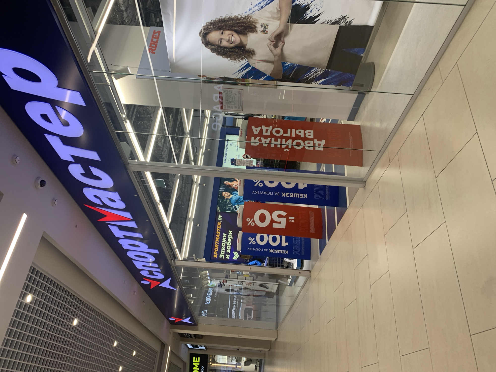 The image shows the exterior of a Sportmaster store in a shopping mall. The store has a large illuminated sign with the Sportmaster logo and name in Russian. In the window, there are posters advertising sales and promotions, including "Cashback up to 100%" and "Double benefit". A large poster on the right features a smiling girl with curly hair. The floor of the mall is tiled, and the lighting is bright. The interior of the store is visible through the glass windows, revealing displays of sporting goods. The location is identified as Hrodna, Belarus, based on the presence of the Sportmaster store chain, which operates in that country. There are no people clearly visible outside the store.