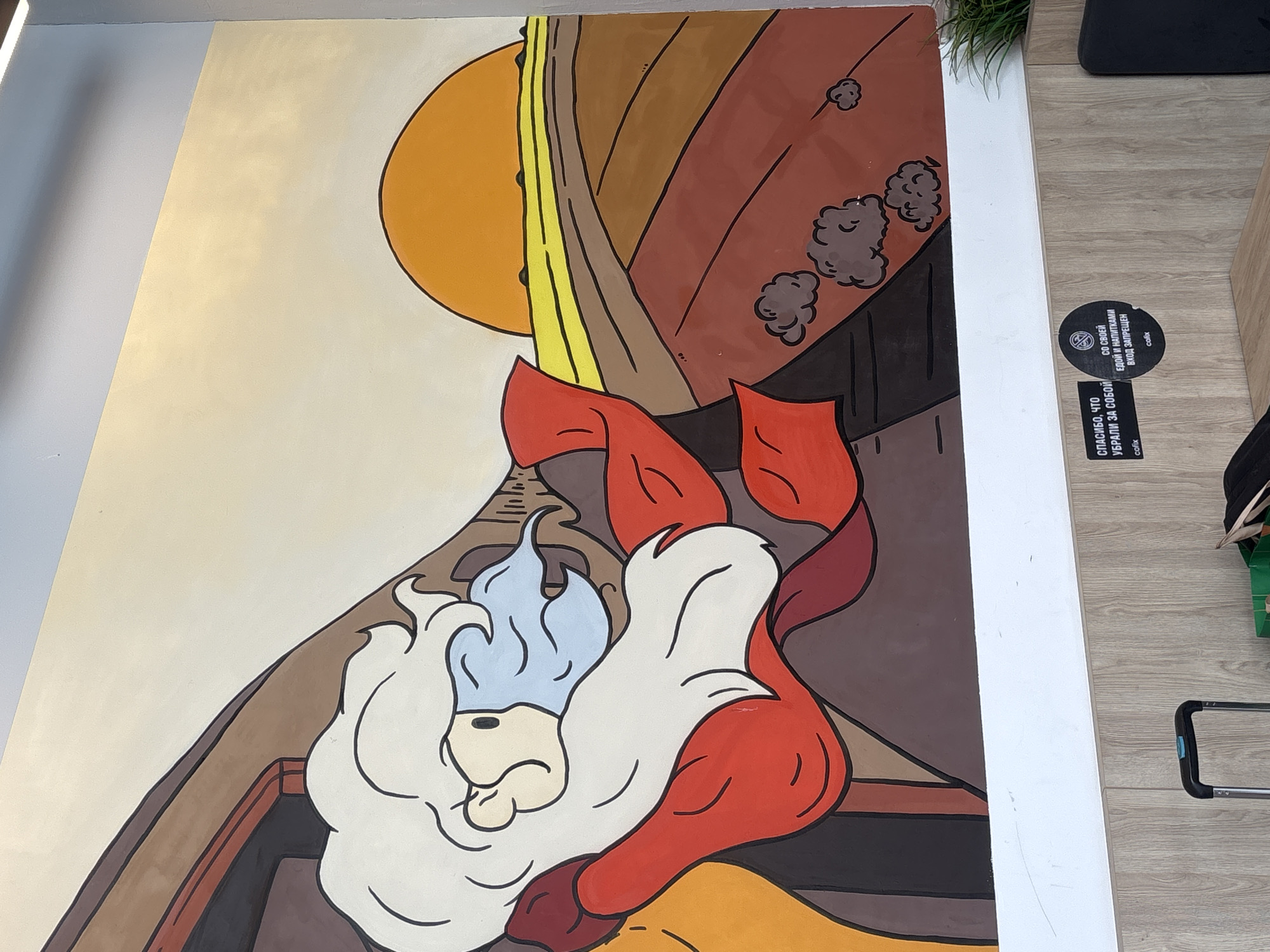 A mural painting on a wall depicts a cartoonish character with long white hair and a red cloak, looking out at a sunrise over a field. The character has blue hair wisps. The field is depicted with various shades of brown and orange, with yellow lines representing the horizon. The sky is a gradient from light yellow to white, with a large orange sun partially visible. The painting is on a wall in a public space, as evidenced by a white shelf below the mural, where two stickers are affixed. One sticker reads "СПАСИБО, ЧТО УБРАЛИ ЗА СОБОЙ" (Thank you for cleaning up after yourself) with "cofix" below it. The other sticker has a no-food-or-drink-from-outside icon and reads "СО СВОЕЙ ЕДОЙ И НАПИТКАМИ ВХОД ЗАПРЕЩЕН" (Entry with your own food and drinks is prohibited) with "cofix" below it. A black suitcase handle is visible on the left, resting on a light brown wooden surface, suggesting the location is a waiting area or a travel hub. A green object, possibly a bag or container, is partially visible on the table in front of the suitcase. The overall scene is indoors, with a bright, artificial light source from above casting subtle shadows. The specific location in Moscow, Russia cannot be determined from the image alone.