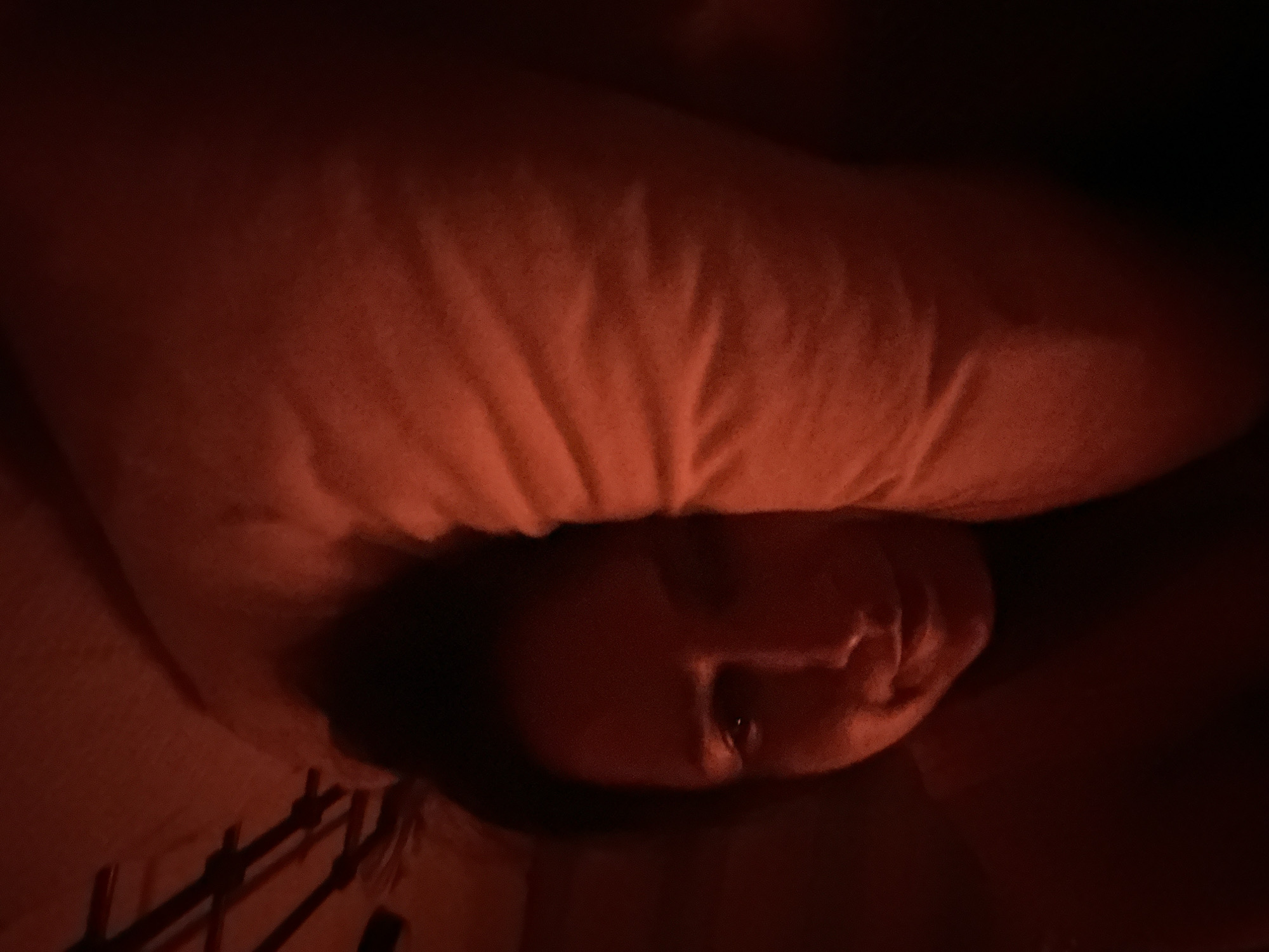 A close-up, low-angle shot depicts a person's face partially obscured by a pillow. The scene is dimly lit with a reddish hue. The person's face is tilted towards the camera. In the lower left corner, part of a wooden structure is visible. The location is not discernible from the image.