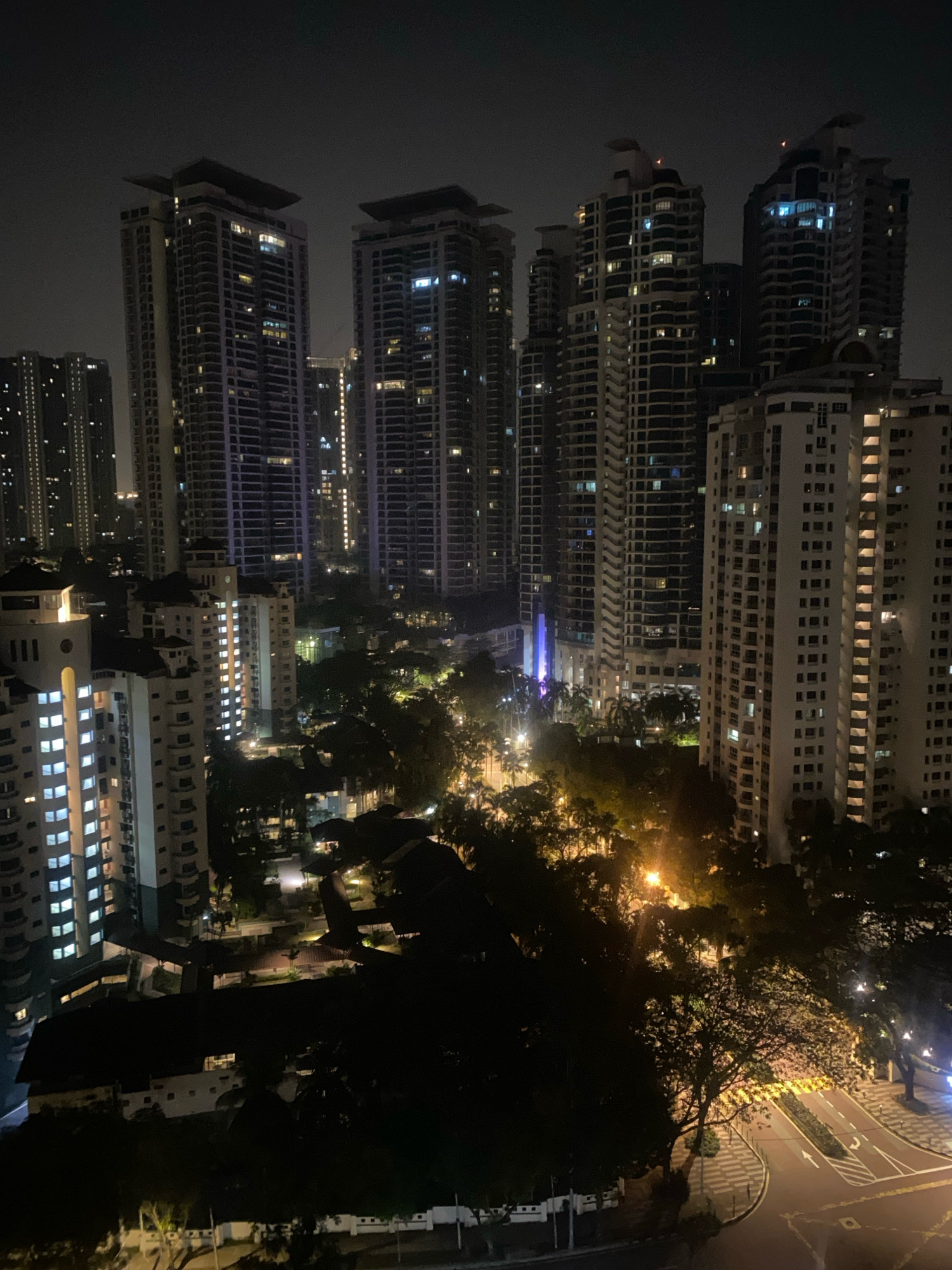 This is a nighttime aerial view of a city landscape in Kuala Lumpur, Malaysia, featuring a cluster of high-rise residential buildings and surrounding greenery. The buildings are illuminated from within, with many windows lit, indicating occupancy. Some of the taller skyscrapers have distinctive architectural features, such as stepped roofs and balconies. The middle ground is filled with trees and lower buildings, some with illuminated windows as well. A road with visible lane markings and streetlights is present in the foreground, suggesting an urban environment with active infrastructure. The sky is dark and overcast, typical of a city at night. There are no visible people or specific actions depicted in the scene.