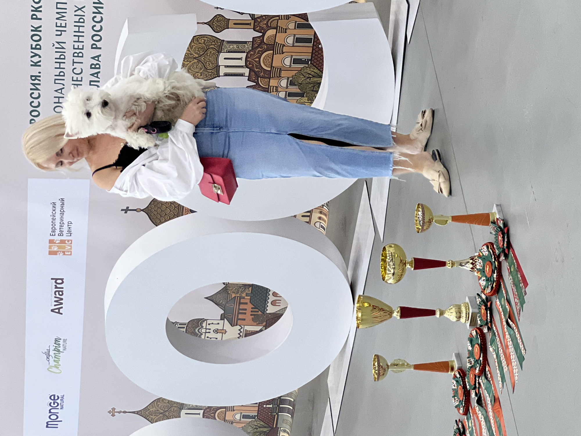 A blonde woman in a white shirt and denim skirt holds a small white dog. She wears beige flats with bows and a red purse. In the background are large white "O" shapes with images of Russian churches. In the foreground are several gold trophies and ribbons. The scene appears to be an outdoor event in Бутынь Днт, Russia.