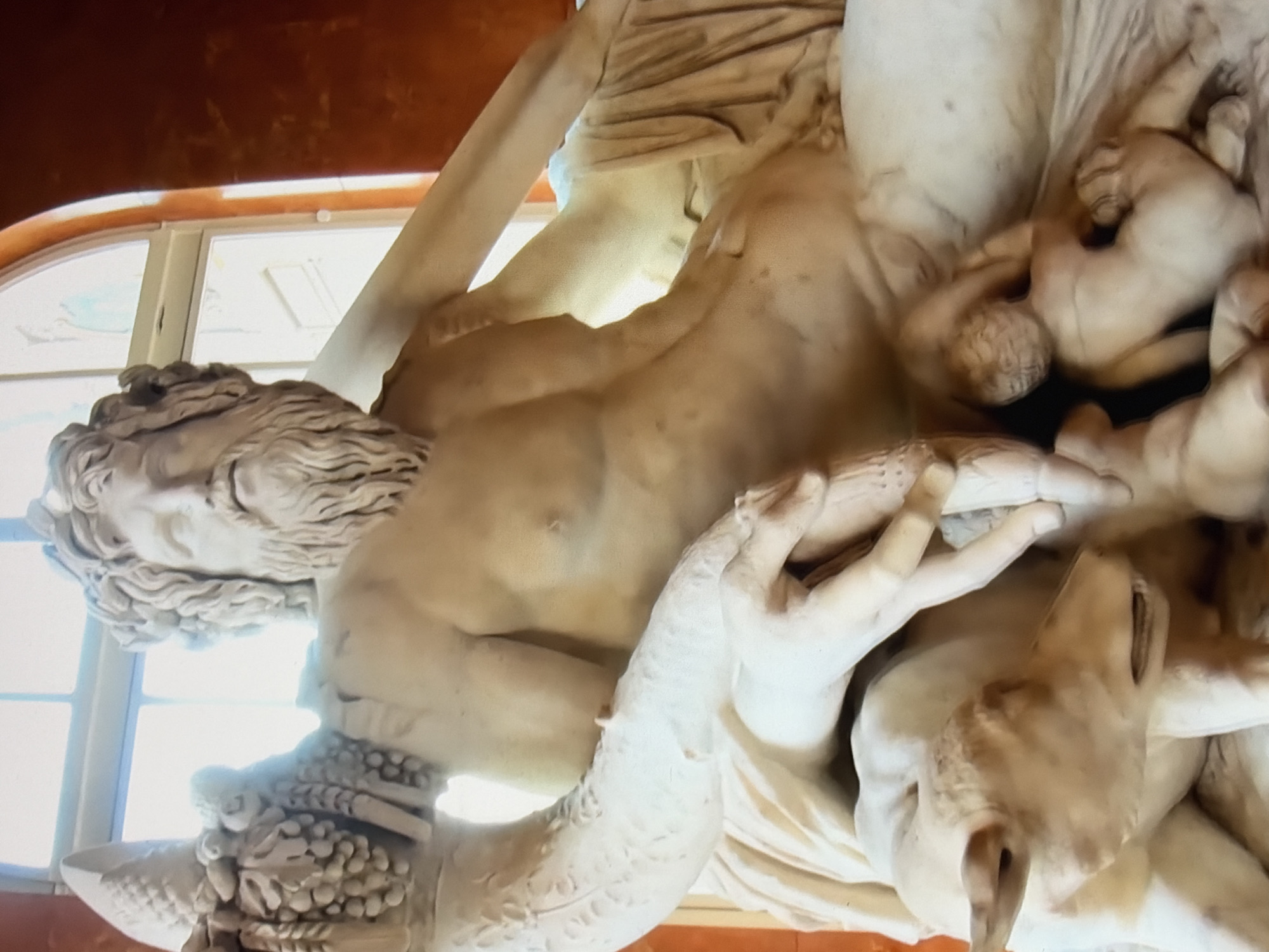 This is a close-up, eye-level photograph of a marble sculpture of a male figure, likely from antiquity, seated and draped in classical robes. The figure has a long, wavy beard and hair, and a muscular physique. His left arm rests on his lap, and his right arm is extended outwards. He is holding an object in his left hand. Below him, two animal figures are depicted: a fawn-like creature to his left, and a smaller, child-like figure or possibly another animal to his right. The sculpture is well-lit, with natural light coming from a large window in the background. The background also shows a reddish-orange wall and a segment of the window frame. The sculpture appears to be located indoors, possibly in a museum or gallery. The image is focused on the central part of the sculpture, emphasizing the details of the figure's face, torso, and the surrounding elements.