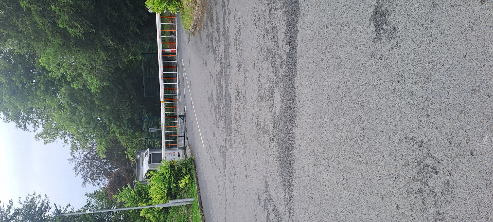 This is a medium shot, eye-level, outdoor, daylight, full frame photograph of the entrance to Breaffy House Resort in Breaffy, Ireland. The scene is a paved road leading to a security barrier. The barrier is a white, horizontal bar with orange stripes, currently lowered and blocking the entrance. To the left of the barrier is a small, gray security hut with a white awning and a sign that reads "BREAFFY HOUSE WELCOME CENTRE". A tall, gray metal pole with a security camera attached stands to the left of the security hut. Lush green trees and foliage line the sides of the road and surround the entrance, with a gray, overcast sky visible above the tree line. The asphalt road surface is wet and appears to have some dark patches, possibly from rain. No people are visible in the frame.