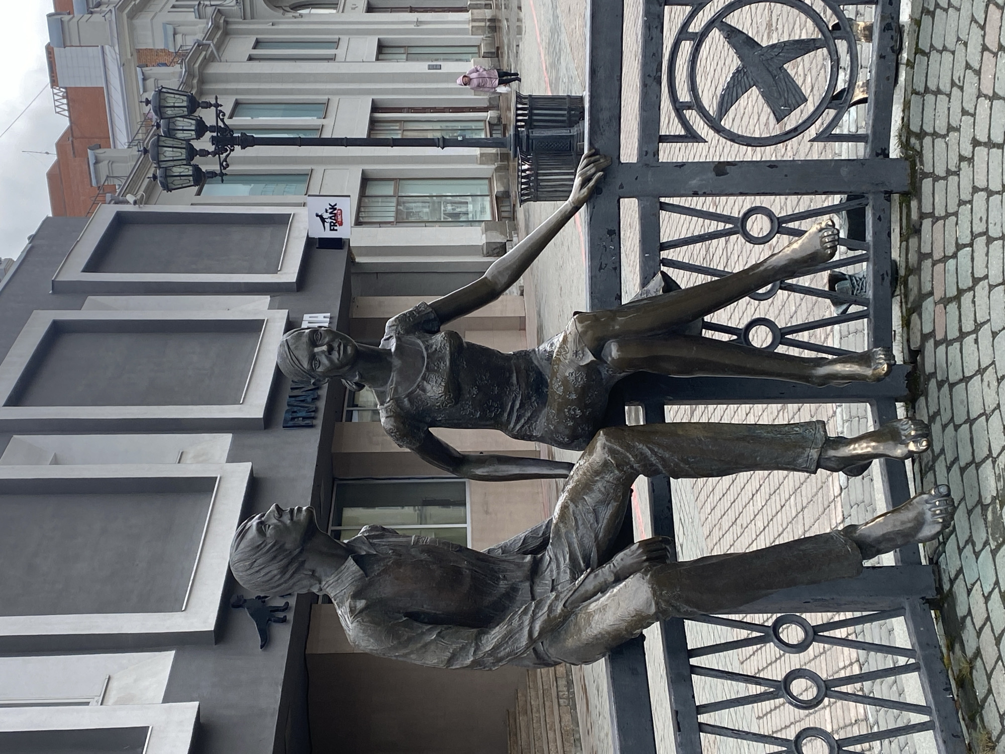 A bronze sculpture of a man and woman is attached to a black metal fence, which is set on a brick sidewalk. The man is lying down, propped up on his arm with his legs extended, and the woman is sitting on his legs with her legs extended and her arm reaching up to the fence. Behind the sculpture is a building with a sign that reads "FRANK." Further back is a building with a streetlamp in front. A person in a pink coat is walking on the sidewalk in the distance. The location is Yekaterinburg, Russia.