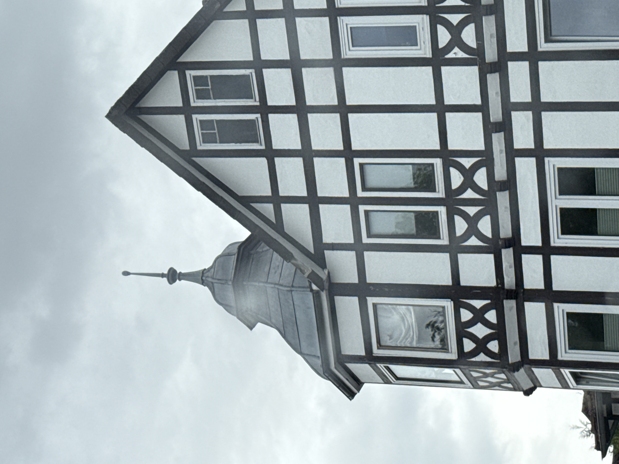 A half-timbered building in Bad Sachsa, Germany, under a cloudy sky, features a prominent pitched roof with dark timbers on a white facade. A round tower with a grey conical roof topped by a spire protrudes from the left side of the building. The building has multiple windows, some with white frames and clear glass, others with dark panes, and decorative "X" shaped patterns between some of the windows on the lower levels. A small section of another building with a darker roof is visible on the far left.