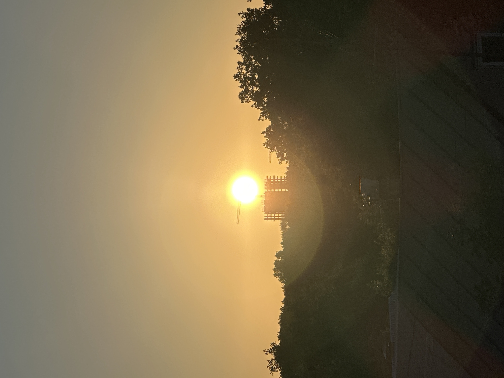 This image captures a sunset scene in Nizhny Novgorod, Russia. The sun, a bright, diffused orb, is positioned in the center of the frame, casting a warm, golden-orange glow across the sky. The sky is a gradient of pale yellow to a muted grey, suggesting the sun is low on the horizon.
In the mid-ground, silhouetted against the sun, is a tall, lattice-like structure, possibly a transmission tower or a part of industrial infrastructure. To the right of the sun and extending towards the foreground is a dense mass of dark green foliage, representing trees and vegetation. The overall impression is one of a serene, natural setting bathed in the soft light of dusk.
In the far right of the image, a dark, sloped surface with horizontal lines is visible, possibly a rooftop or an inclined plane. A small, rectangular dark opening, likely a window or vent, is also present on this surface. The contrast between the bright sky and the dark silhouettes of the structures and trees emphasizes the dramatic lighting of the sunset. The image appears to be taken from an elevated position, looking out over a landscape. The time of day is clearly sunset, and the weather appears clear, allowing for an unobstructed view of the sun. No text is visible in the image.