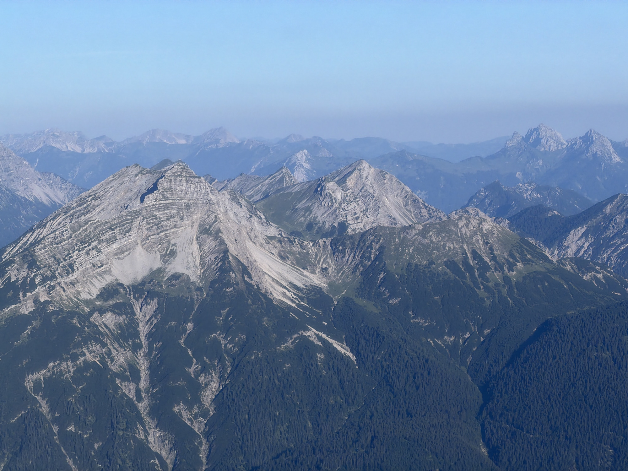 This is a landscape photograph featuring a panoramic view of a mountain range under a clear blue sky. The foreground is dominated by a large, rugged mountain with steep, rocky slopes and dense evergreen forests covering its lower sections. Streaks of lighter rock and scree are visible on the upper slopes, indicating a weathered and eroded terrain.
Further back, several layers of mountains recede into the distance. These mountains appear progressively softer and more muted in color due to atmospheric perspective, fading into shades of blue and gray. Peaks vary in shape, from sharp, jagged summits to more rounded, undulating ridges. The overall impression is one of vastness and natural grandeur.
The location is identified as the area around Grainau, Germany, a municipality situated in the Bavarian Alps. The scene captures the typical alpine environment of this region, characterized by its dramatic mountain formations and extensive forests. There are no people or man-made structures visible in the image, emphasizing the wild and natural state of the landscape. The lighting suggests it is daytime, likely morning or afternoon, with the sun illuminating the mountains.