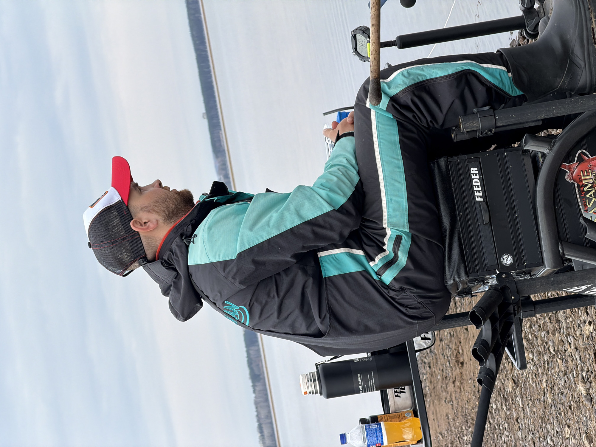 A man is seated on a fishing chair by a body of water, possibly the Kama River, in the city of Chaikovsky, Perm Krai, Russia. He is wearing a black and turquoise jacket, black pants with turquoise stripes, and black rubber boots. He also wears a red, white, and black baseball cap. He is looking upwards.
The chair is equipped with a "FEEDER" box, fishing rod holders, and a side table. On the table, there is a thermos, a box, and bottles of liquid. A fishing rod is visible.
The background consists of water and a distant, tree-lined shore under a pale sky. The foreground shows a pebbly shoreline.