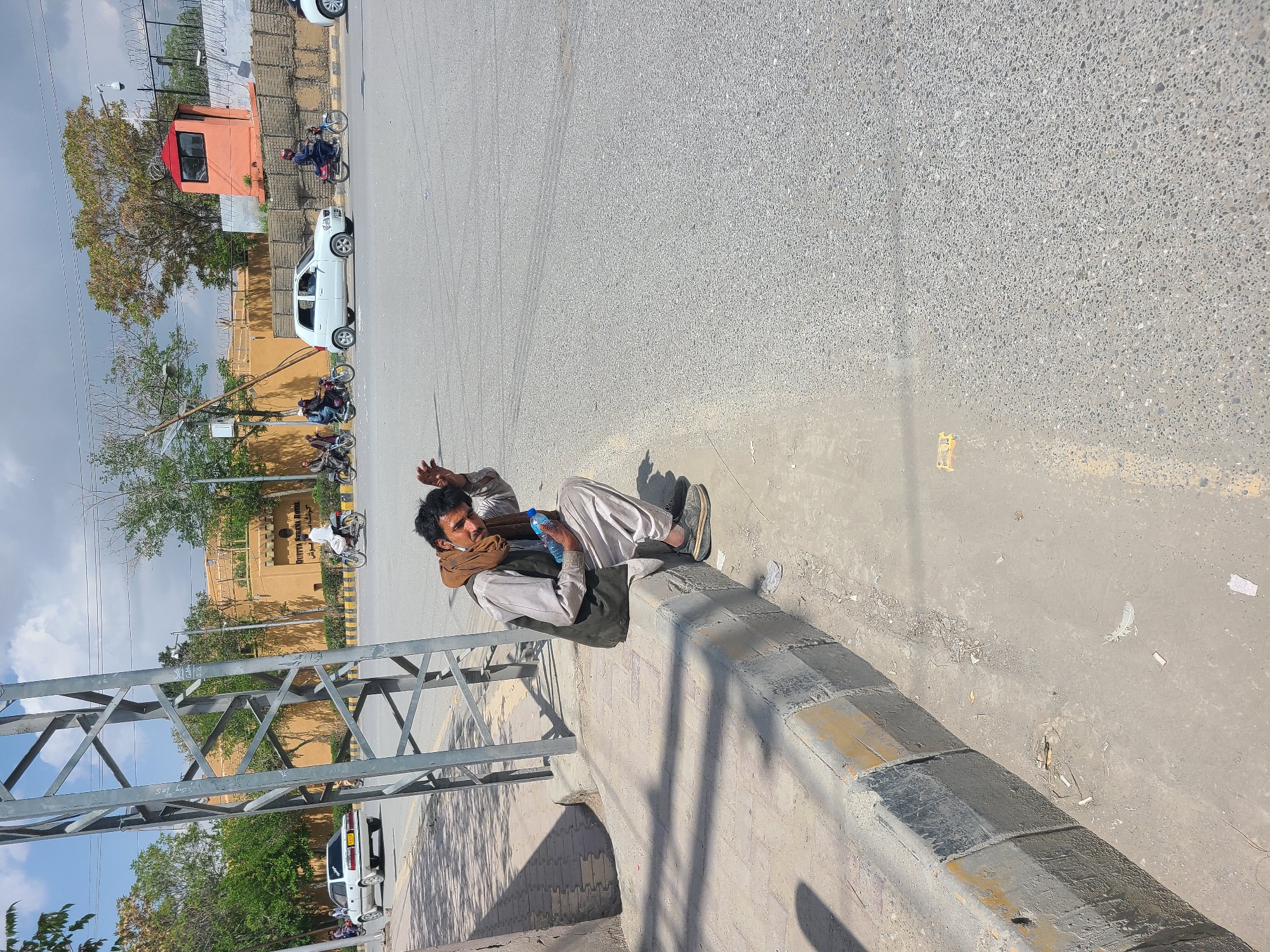 A man in traditional Pakistani clothing is lying on a concrete curb in an urban environment in Quetta City Tehsil, Pakistan. He is holding a water bottle. In the background, there is a street with cars and motorcycles. There is also a building and a metal structure visible. The weather appears to be sunny.