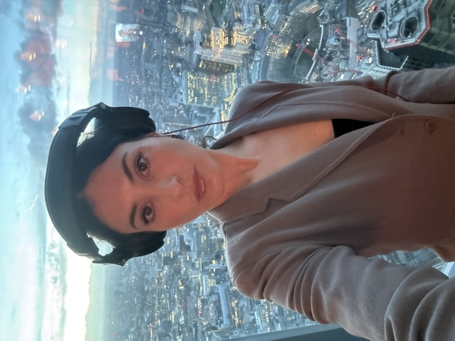 A female individual is depicted in a waist-up, forward-facing pose, taking a selfie indoors. She possesses dark hair and dark eyes, and is wearing large, black over-ear headphones connected by a thin red cable descending toward her chest. Her attire comprises a light beige or brown blazer worn over a dark, likely black, inner garment.
The background consists of a panoramic view of the London cityscape at dusk, observed through a large window. The sky exhibits a blue-grey tone with cloud formations. Numerous commercial buildings are visible below, many with interior lights illuminated. The distant London Eye is identifiable, confirming the location as London, United Kingdom, specifically from an elevated position within the City of London district. Reflections of indoor lights and a faint image of a figure on what appears to be a screen are visible on the window's surface to the right of the subject.