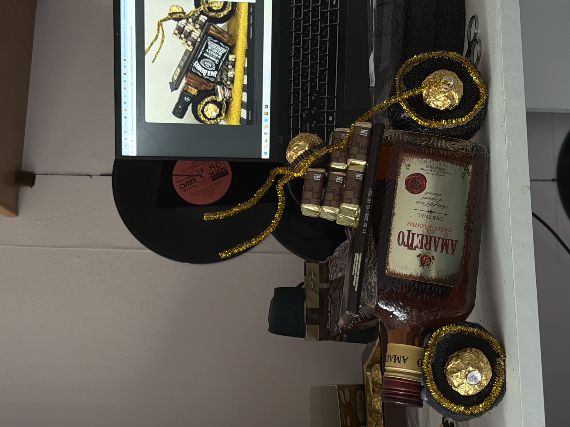 The image displays an indoor scene, likely a desk or shelf, filled with various objects, suggesting a prepared gift or collection of treats. On the left side, a laptop with an open monitor is visible. The monitor screen shows an image of a decorative arrangement featuring a prominent bottle of "JACK DANIEL'S" whiskey, adorned with gold tinsel and checkered brown and white patterns, possibly from candy wrappers or fabric.
On the desk to the right of the monitor, a black vinyl record, marked "DMM" and "Weerberlig," is partially visible underneath other items. Dominating the foreground is a bottle of "AMARETTO San Remo ORIGINAL" liqueur, featuring a red wax seal and labeled with "Delightful taste of natural almonds," "0.5 L," and "alc 20% vol." The Russian text "ЛИКЕР ДЕСЕРТНЫЙ АМАРЕТТО САН РЕМО ОРИДЖИНАЛ" is also printed on the label.
Arranged beside the Amaretto bottle are several individual chocolate bars, some wrapped, displaying the "EAC" conformity mark and the text "Птичье молоко" (Ptichye Moloko). Dates "11.02.2026 / 05.08.2027" are visible on one of the chocolate packaging. A long strand of gold tinsel string weaves through the items, connecting to two gold-wrapped candies, clearly identifiable as "FERRERO ROCHER," which are set on small black mesh-like bases also decorated with gold tinsel. A pair of scissors is visible tucked behind these items on the far right. The overall arrangement suggests a thoughtful presentation or a collection for personal enjoyment.