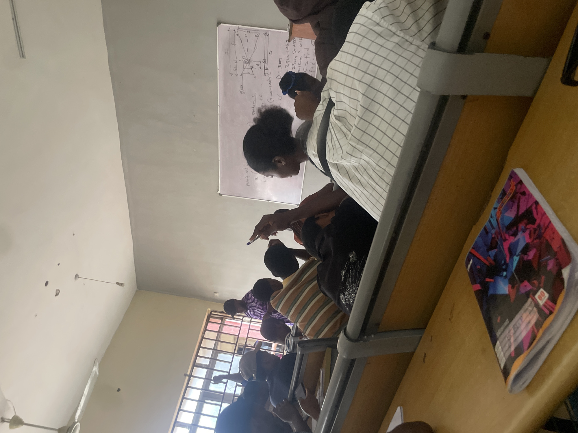 The image depicts a classroom scene in Ihiagwa, Nigeria. The room appears to be a lecture hall with tiered wooden desks arranged in rows. Several students are seated at the desks, facing forward. Some are looking at a whiteboard at the front of the room, which has diagrams and text written on it. Others are engaged with their phones or looking down at their notebooks. A few individuals are standing or walking in the aisles. The room is lit by natural light from a large window on the left side and artificial lighting from overhead fixtures. A ceiling fan is visible on the ceiling. The overall atmosphere suggests an academic setting.
