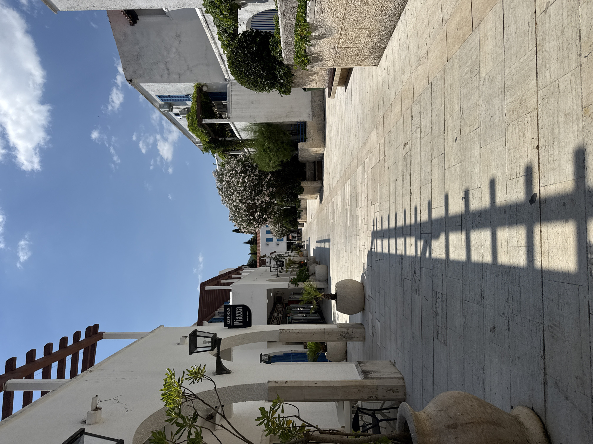 The media file depicts a picturesque street scene in Budva, Montenegro, under a clear blue sky with scattered white clouds. The perspective is from street level, looking down a pedestrian walkway paved with light-colored stone tiles. On the left side of the walkway, there are white buildings with architectural elements like arched doorways, balconies adorned with greenery, and traditional light fixtures. A prominent sign reads "RESTORAN PIZZA".
On the right side of the walkway, a stone wall is visible, casting long, geometric shadows from an unseen fence or structure. The shadows suggest the time of day is likely late afternoon or early morning, when the sun is at a lower angle. Large, rounded stone planters with small palm trees are strategically placed along the walkway, adding to the Mediterranean ambiance.
The scene is tranquil and suggests a pleasant day for a stroll or dining at one of the establishments. The whitewashed buildings, lush vegetation, and paved walkway evoke a sense of charm and vacation atmosphere typical of coastal towns. No people are visible in the frame, emphasizing the peacefulness of the moment. The overall impression is one of a serene and inviting European street.