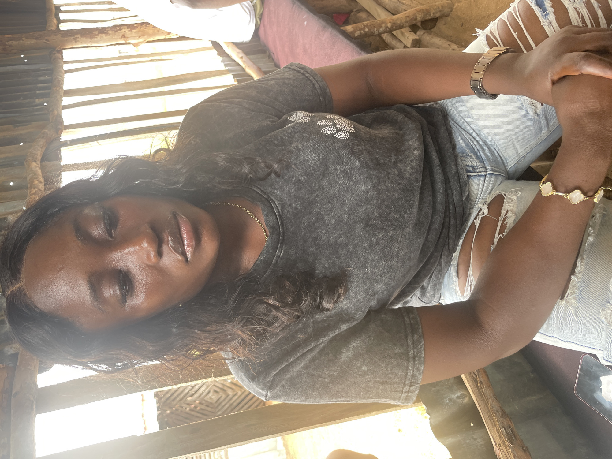 A medium shot captures a Black woman from the chest down, seated and looking downward with her head bowed. She has dark, wavy hair cascading over her shoulders. She is wearing a dark gray, acid-wash t-shirt with a white paw print graphic on the left chest. Her light-wash denim jeans are ripped at the thighs, revealing white fabric underneath. On her left wrist, she wears a gold bracelet with white clover-shaped charms. On her right wrist, she has a metallic watch with a link band. She is seated on a bench with a red cushioned seat, supported by rustic wooden legs. The background is composed of vertical wooden slats, some appearing weathered and others more uniform. Sunlight streams through the gaps in the slats, creating strong highlights and shadows. A portion of what appears to be a corrugated metal roof is visible at the top. The overall environment suggests an outdoor, possibly rustic setting in Durumi, Nigeria.