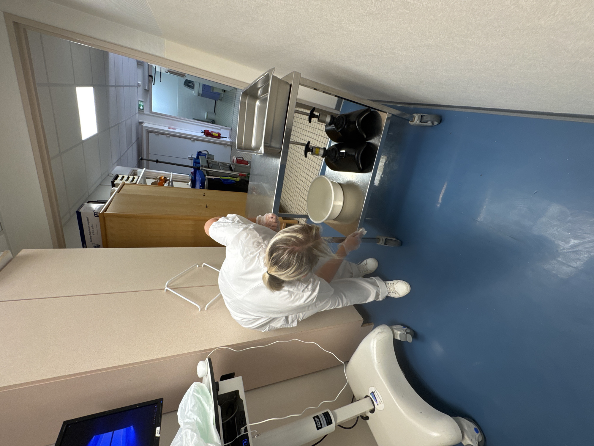A person in a white uniform and gloves is cleaning a stainless steel cart in what appears to be a medical facility. The cart has two shelves. The top shelf holds a metal basin, and the bottom shelf holds two black pump bottles and a white bowl. The floor is blue. A medical monitor on a stand is visible to the left of the cart, and a light wood cabinet is to the right. In the background, a hallway with tiled walls and doors can be seen, with a fire extinguisher mounted on the wall. This scene could be located in Tourcoing, France, based on the provided context, and is likely within a hospital or clinic setting.