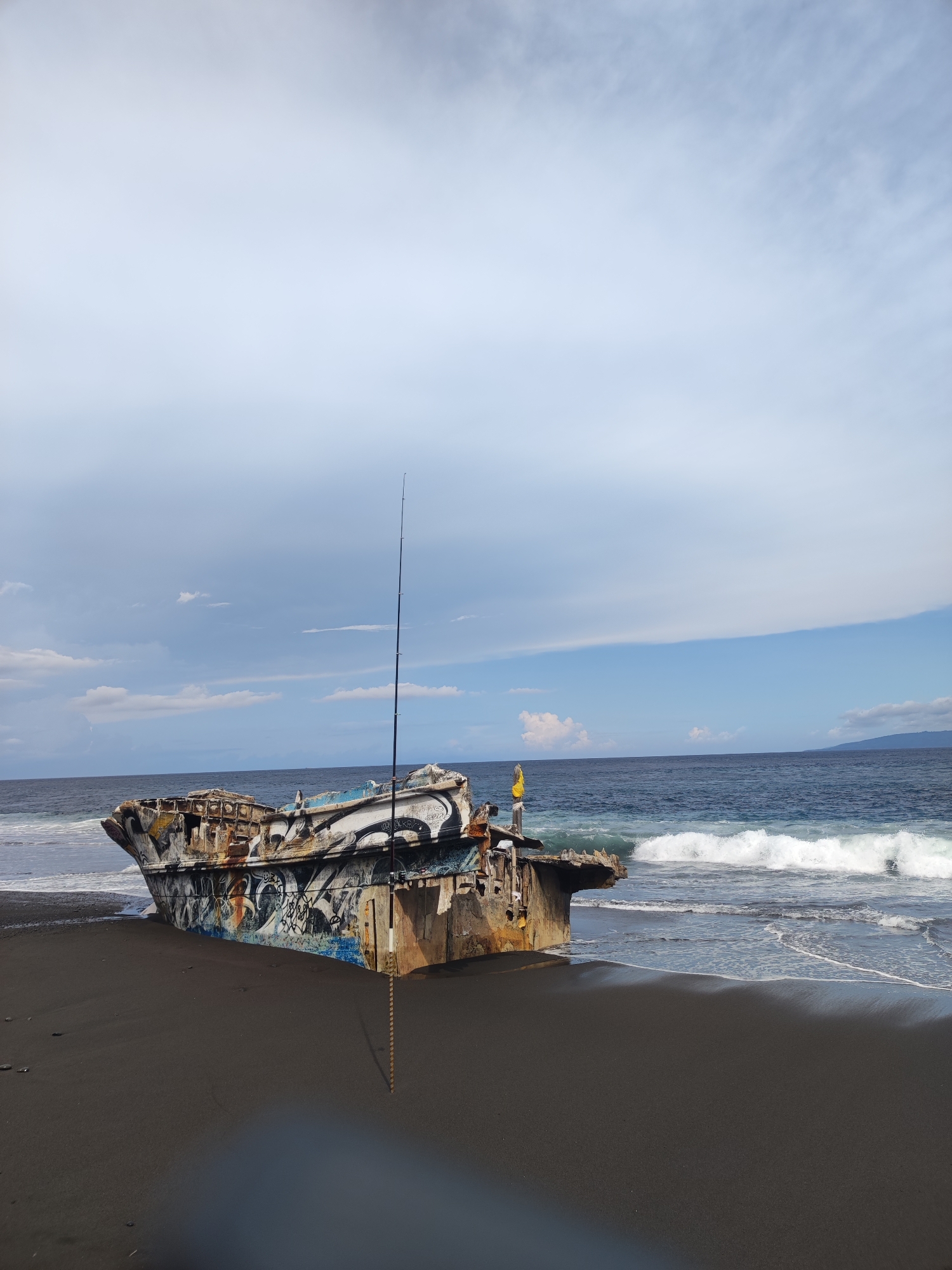 The image shows a beached, heavily graffitied wreck of a ship on a black sand beach. The ship is rusted and partially submerged in the sand, with waves lapping at its side. A fishing rod is propped up near the ship, its line running towards the water. A person wearing a yellow top is standing on the wreck. The ocean is in the background with a distant landmass visible. The sky is partly cloudy. The scene is located on a beach in or near Padangbai, Indonesia.
