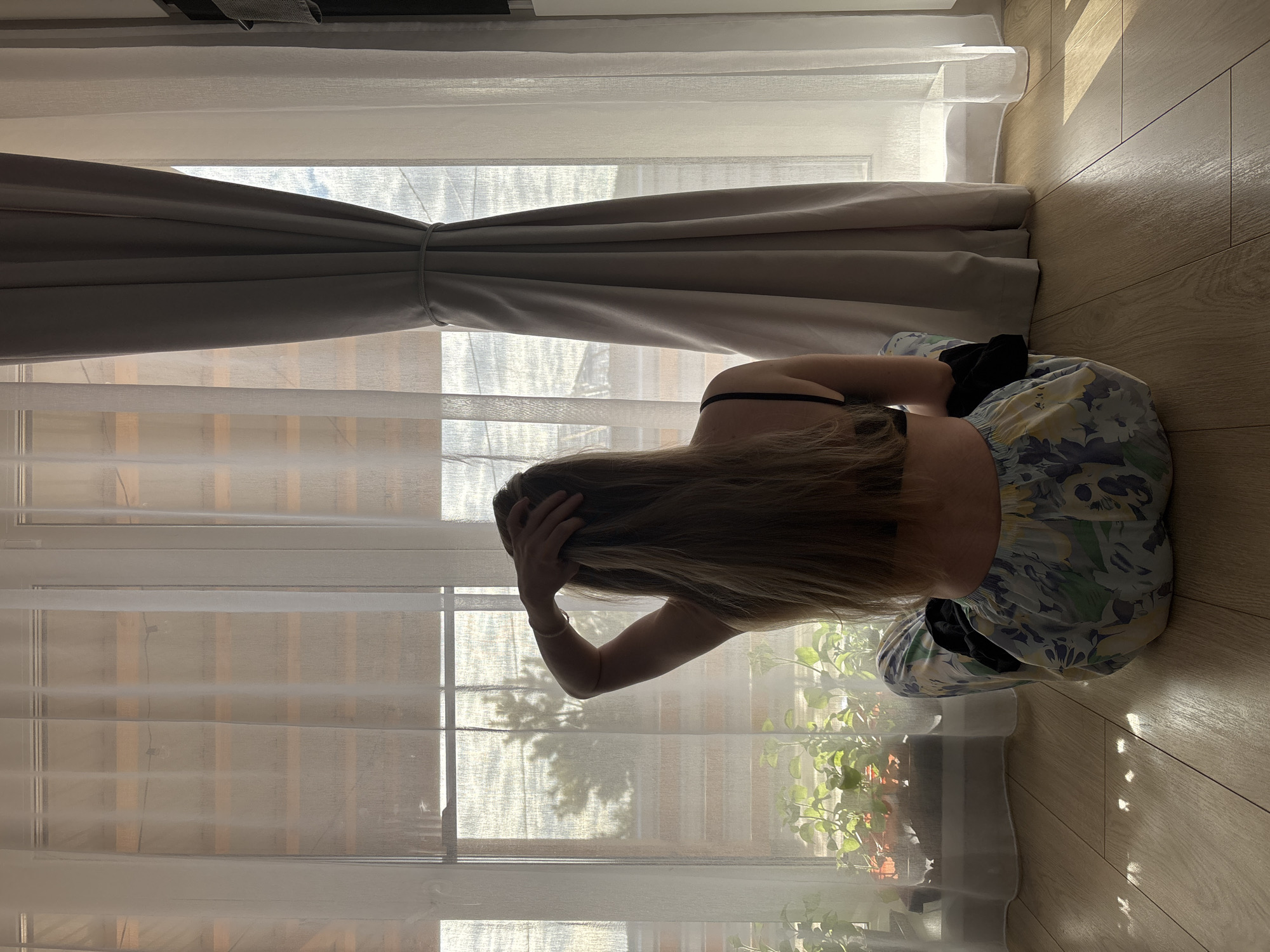 A woman with long blonde hair is sitting on a wooden floor, facing a window. She is wearing a floral dress and black socks. Her left hand is raised, touching her head. Behind her is a large window covered with white, sheer curtains and a grey drape tied in the middle. Outside the window, greenery is visible. The setting is indoors, with the light suggesting it is daytime. The location is Chaikovsky, Russia.