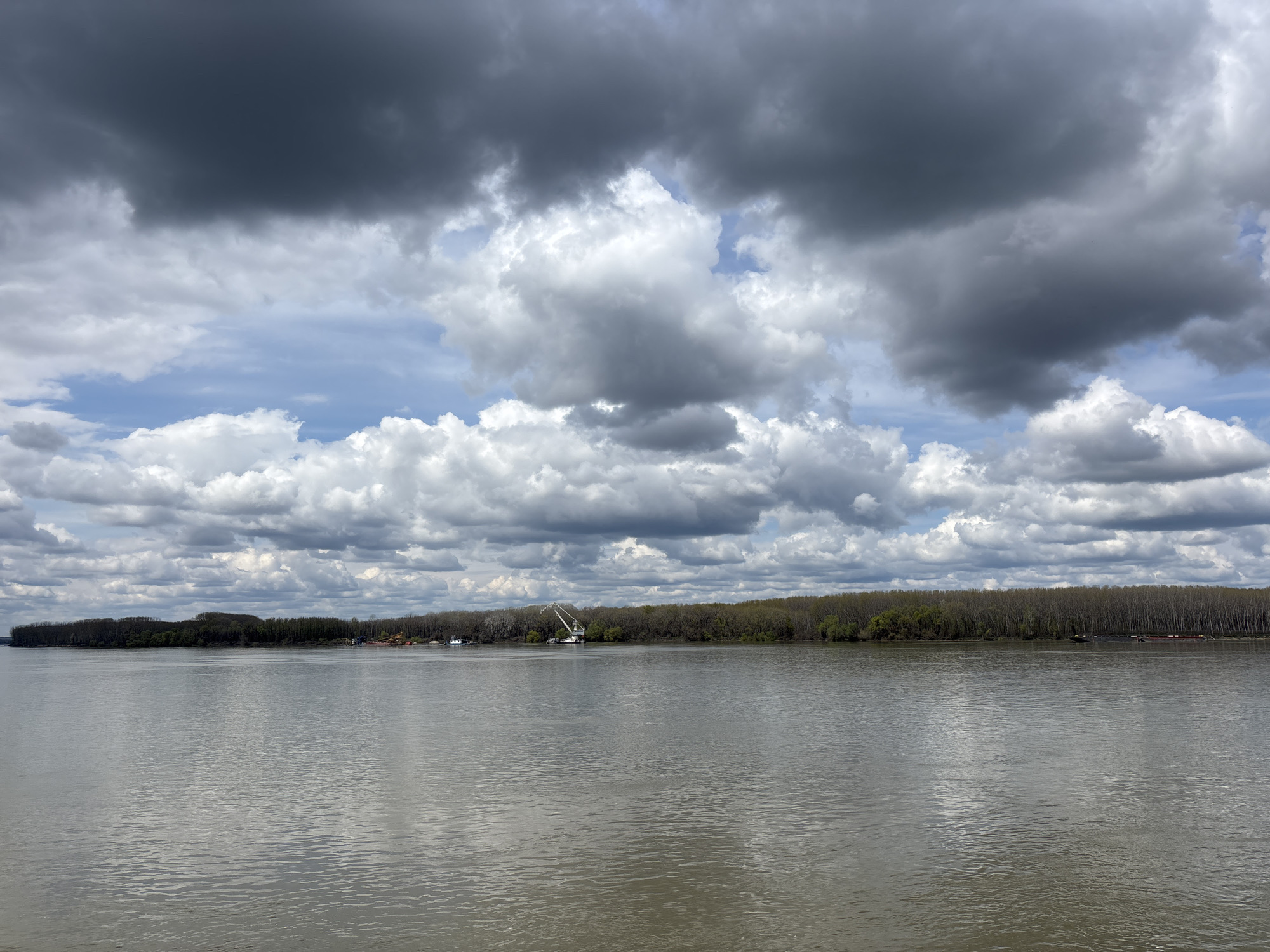 The image depicts a wide river under a cloudy sky. The clouds are a mix of dark, ominous formations and lighter, scattered ones. A line of trees borders the far side of the river, appearing as a dark strip against the horizon. Near the center of the horizon line, a crane-like structure is visible, possibly a piece of industrial equipment. The river surface is mostly calm, reflecting the sky above. Given the context of Silistra, Bulgaria, this is likely a view of the Danube River. There are no people visible in the frame. The overall scene suggests a somewhat overcast day in an industrial or natural area along the riverbank.