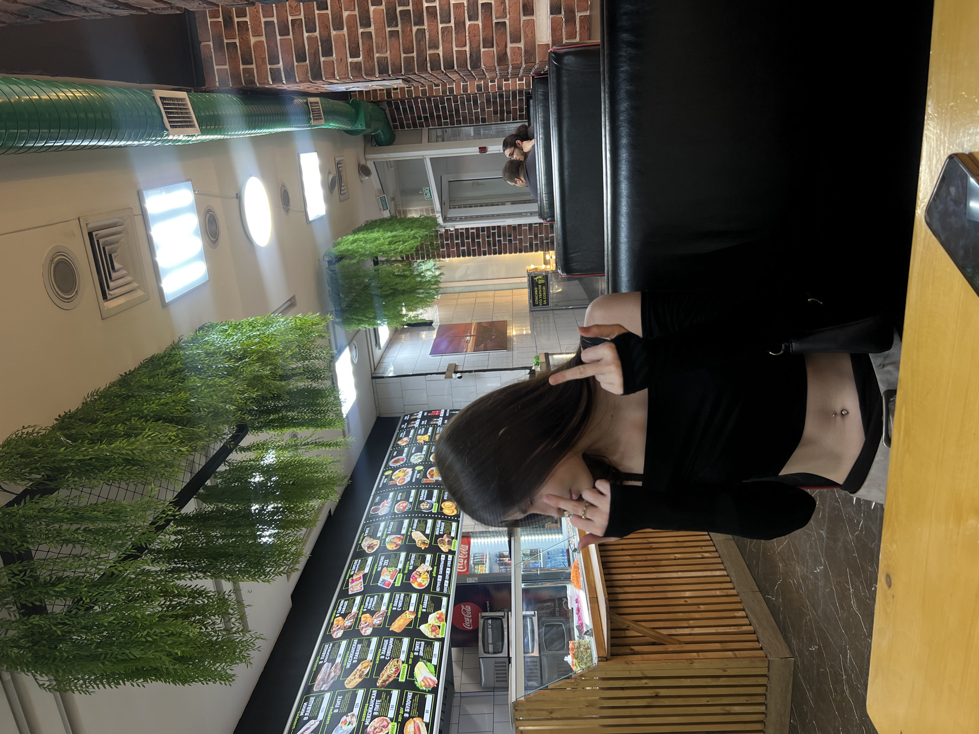 In a fast-food establishment in Saint Petersburg, Russia, a young woman with long dark hair, wearing a black off-the-shoulder top and light-colored pants, is seated at a table. She is looking to her left and is holding up both middle fingers in an offensive gesture. A black purse is on the seat next to her. In the background, a menu board displays various food items, and a display case shows food preparations. Other patrons are seated at tables in the background. The environment features brick walls, hanging artificial plants, and fluorescent lighting.