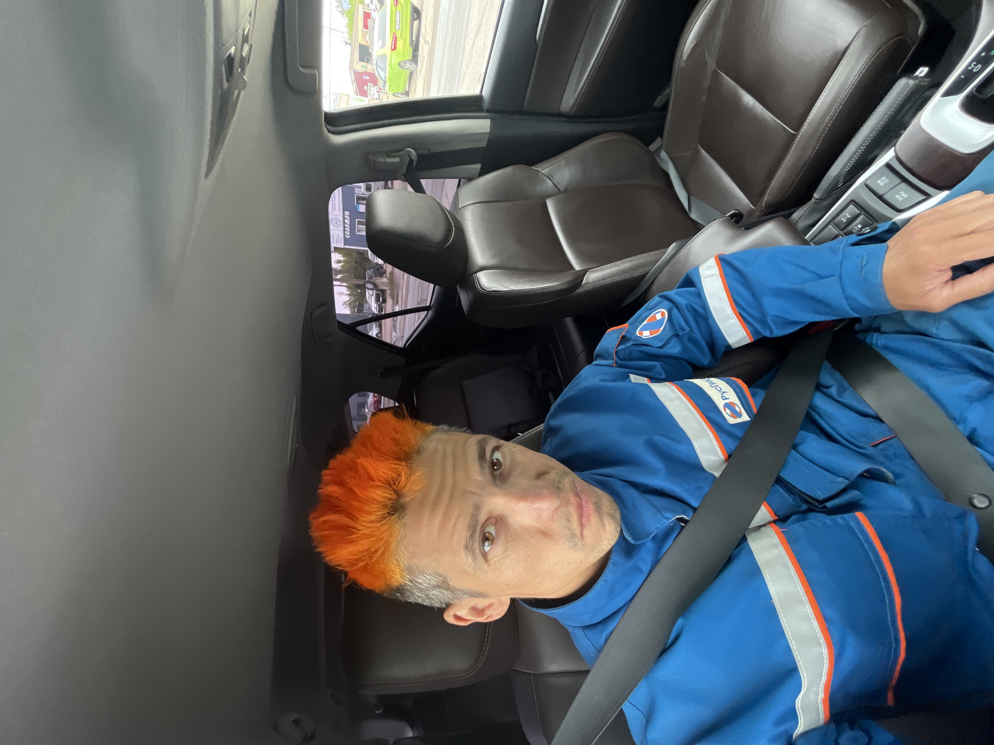 The image captures a man with striking bright orange hair and visible grey roots, seated in the driver's seat of a car. He is wearing a blue uniform with reflective white and orange stripes across the chest and arms. A logo on his left arm reads "РУССКИЙ ПРОДУКТ" (Russian Product) in white text on a blue background. He is wearing a seatbelt and looks directly at the camera with a neutral to slightly inquisitive expression.
The car's interior features brown leather seats, a grey headliner, and a center console visible to the man's right, showing gear selector and buttons like "ECO MODE" and "PWR MODE." Sunlight streams into the vehicle, indicating it is daytime.
Through the car's windows, an outdoor scene is visible. A light green vehicle is parked further away, and a building with Cyrillic text is discernable. The text on the building clearly reads "СЛАДОСТИ" (Sweets/Confectionery). The overall setting appears to be an urban or commercial area in Chaikovsky, Russia, consistent with the visible Cyrillic text. The image is oriented sideways, suggesting it was taken from a smartphone held vertically.