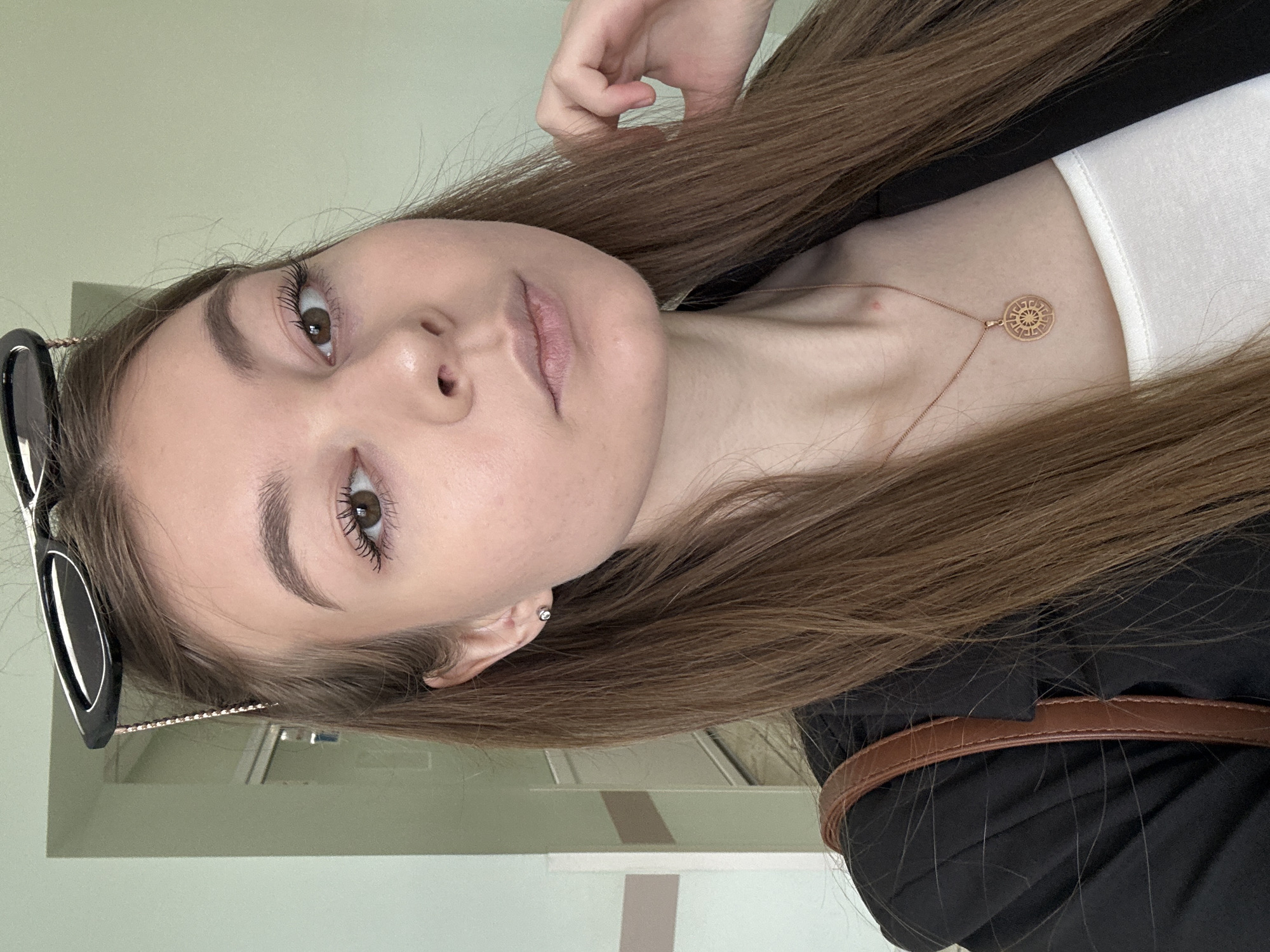 The image presents a close-up portrait of a young woman, slightly angled. She has long, straight brown hair, fair skin, and brown eyes. Her eyes are accentuated with mascara, and her eyebrows are neatly groomed. Her lips show a subtle, natural tint.
She is dressed in a white top visible beneath a dark, likely black, blazer or jacket. A delicate gold chain necklace with a circular, ornate gold pendant, possibly depicting a sun-like emblem, rests on her collarbone. A small silver stud earring is visible in her right ear. Black-framed sunglasses are pushed up onto her head, resting in her hair. Her right hand is raised, with fingers gently touching her hair near the top of her head. A brown strap, possibly from a bag or a belt, is visible over her right shoulder/back.
The background is a plain, light green or beige wall, suggesting an indoor setting. Faint architectural lines, possibly a door frame or wall partition, are discernible in the background. The woman looks directly at the camera with a calm, neutral expression. There is no visible text in the image.
