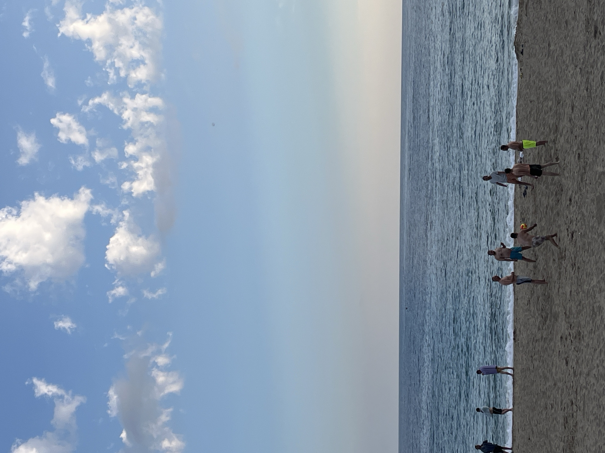 The image depicts a beach scene in Alanya, Turkey. The sky transitions from blue with scattered clouds to a clear, pale blue. Adjacent to the sky is the ocean, with visible waves. Along the shoreline, there's a sandy beach where several people are present. Some individuals are engaged in a game involving a ball, possibly volleyball or football, while others are standing or walking along the shore. The overall setting suggests a typical beachside environment.